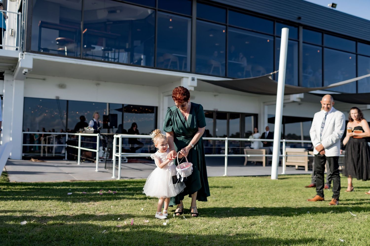 Small child in white dress walks across grass at outdoor wedding venue with modern building.