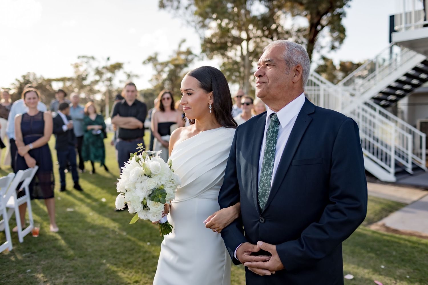 A wedding ceremony processional on a grassy lawn with white flowers at sunset.