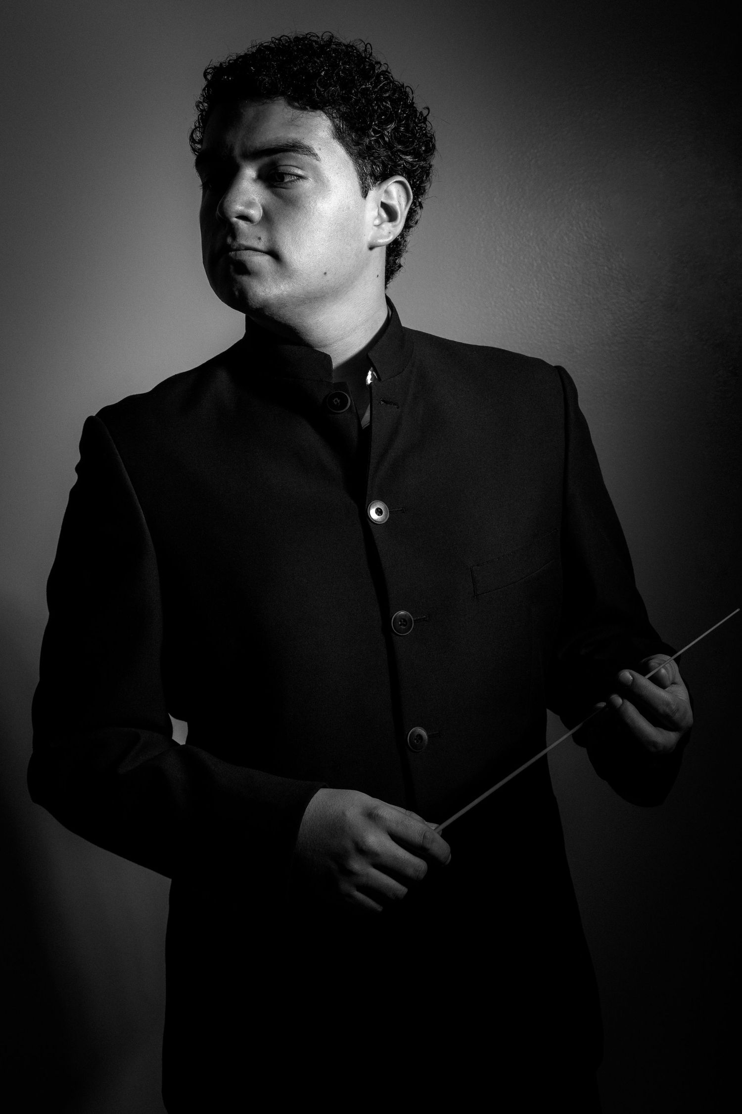 Dramatic portrait of conductor holding baton in formal black jacket against dark background.