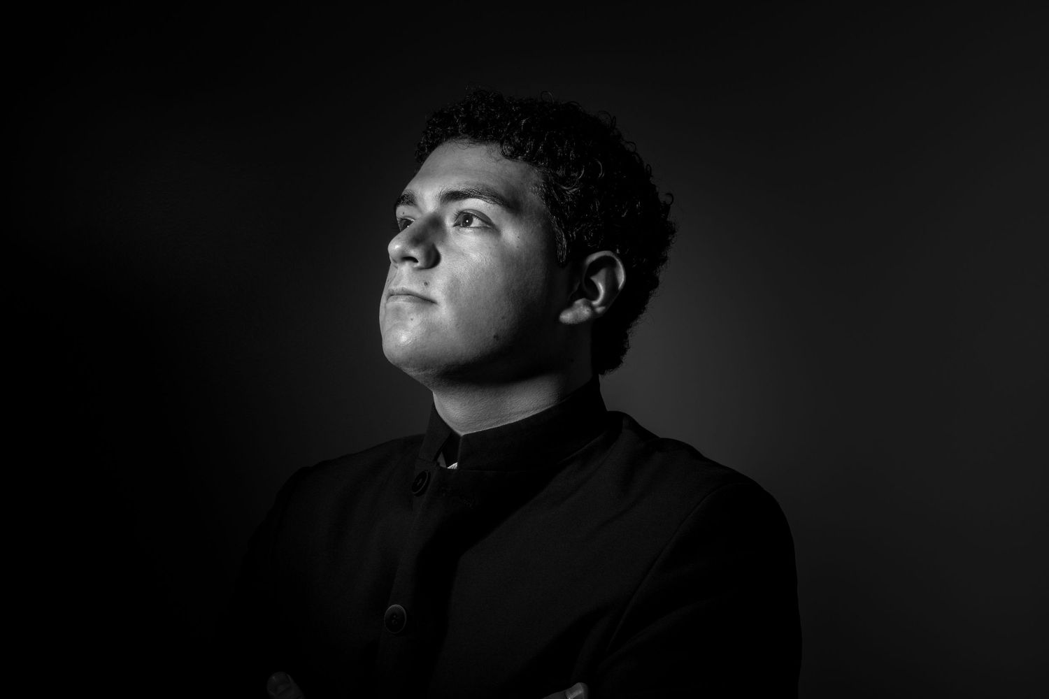 Low-key black and white portrait of conductor looking upward in dramatic lighting.