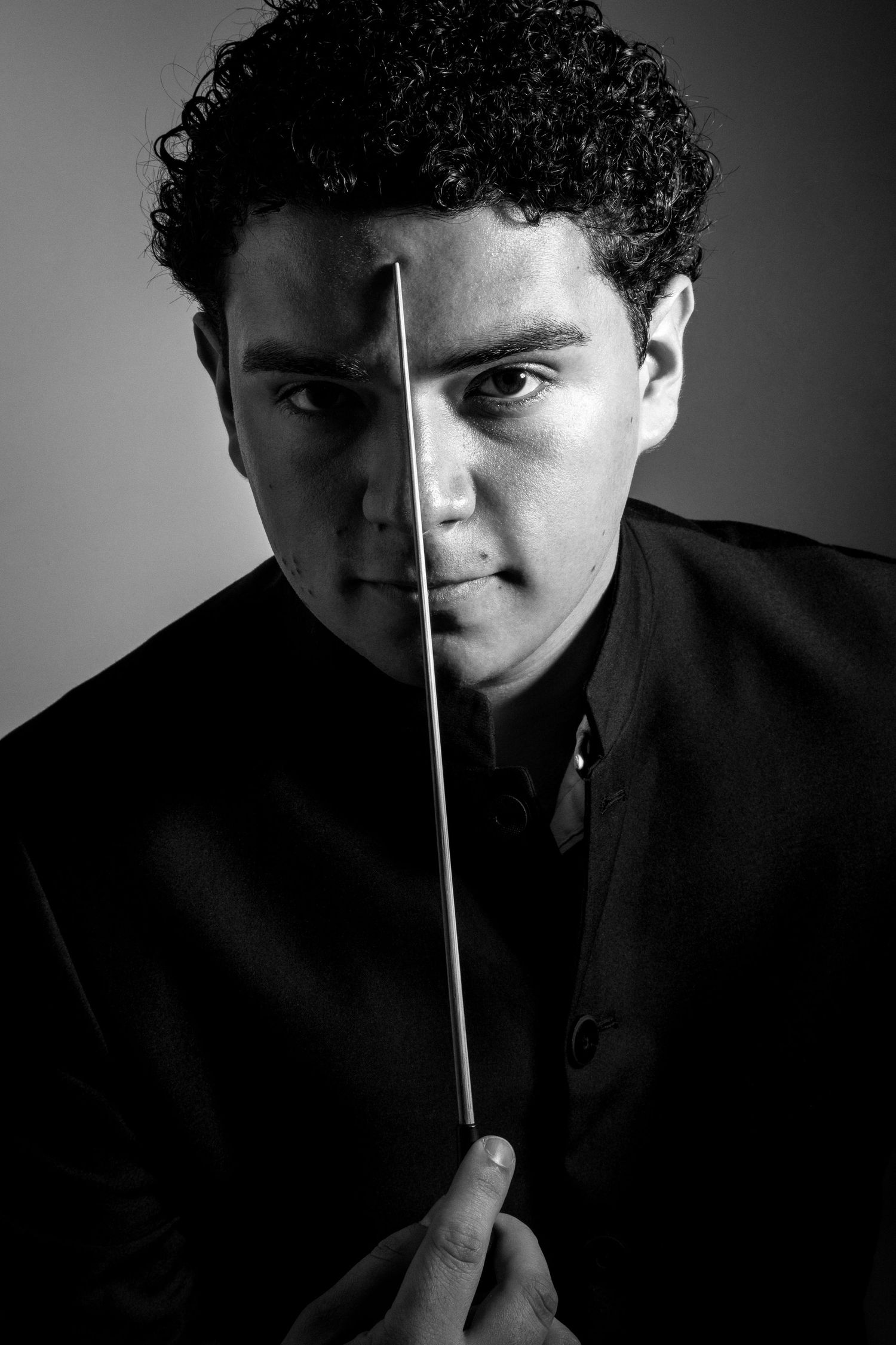 Dramatic black and white portrait of conductor holding baton vertically across face.
