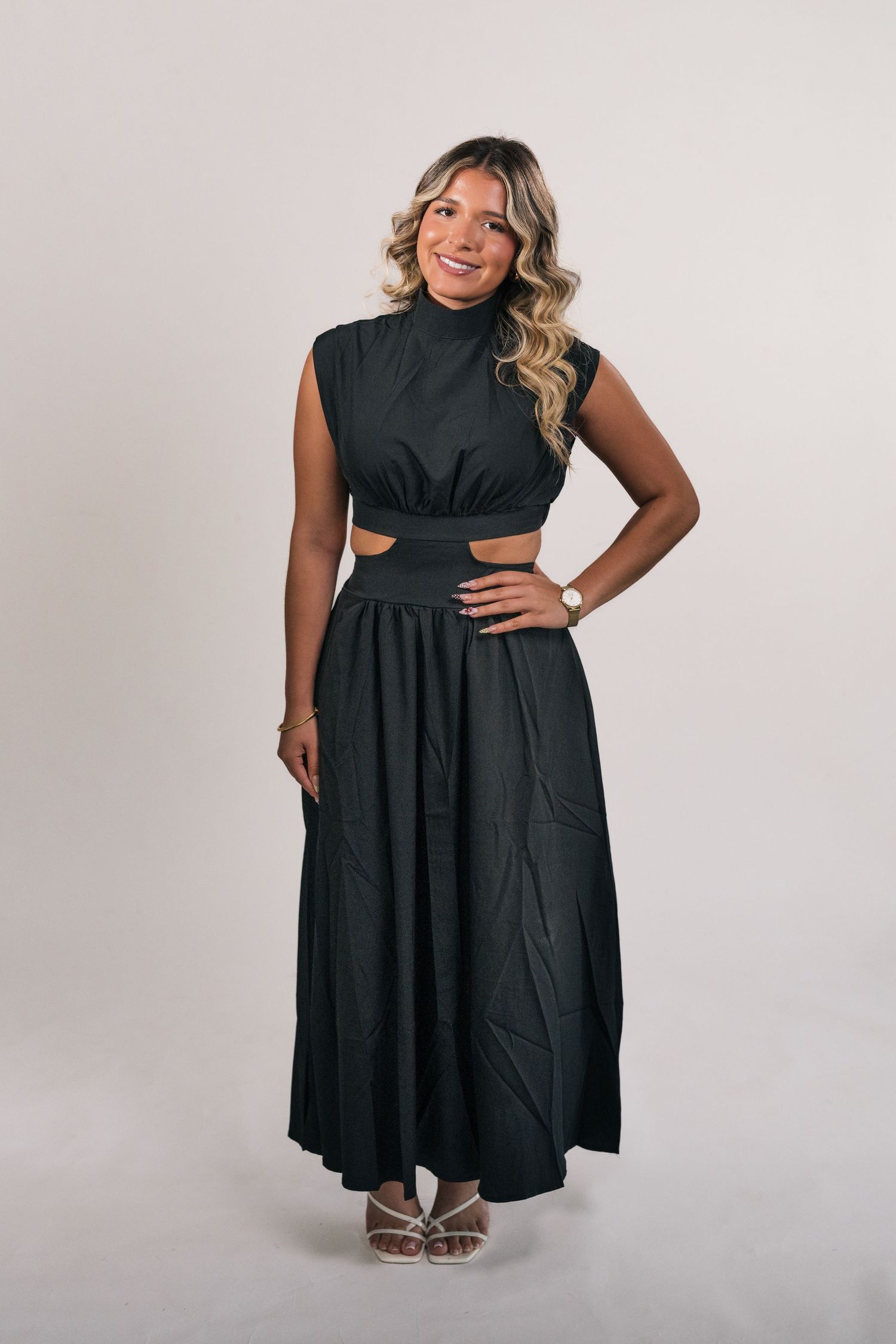Woman wearing a black sleeveless midi dress with cutout waist detail and flowing skirt, paired with white heeled sandals.