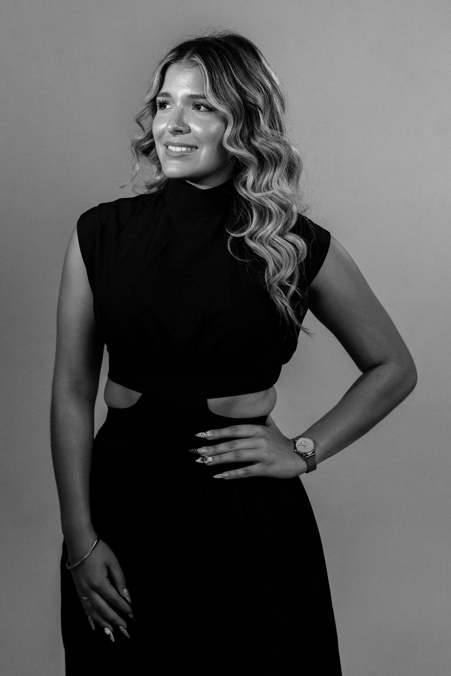 Black and white portrait of a smiling woman in a black sleeveless dress with cutouts, posing with hand on hip.