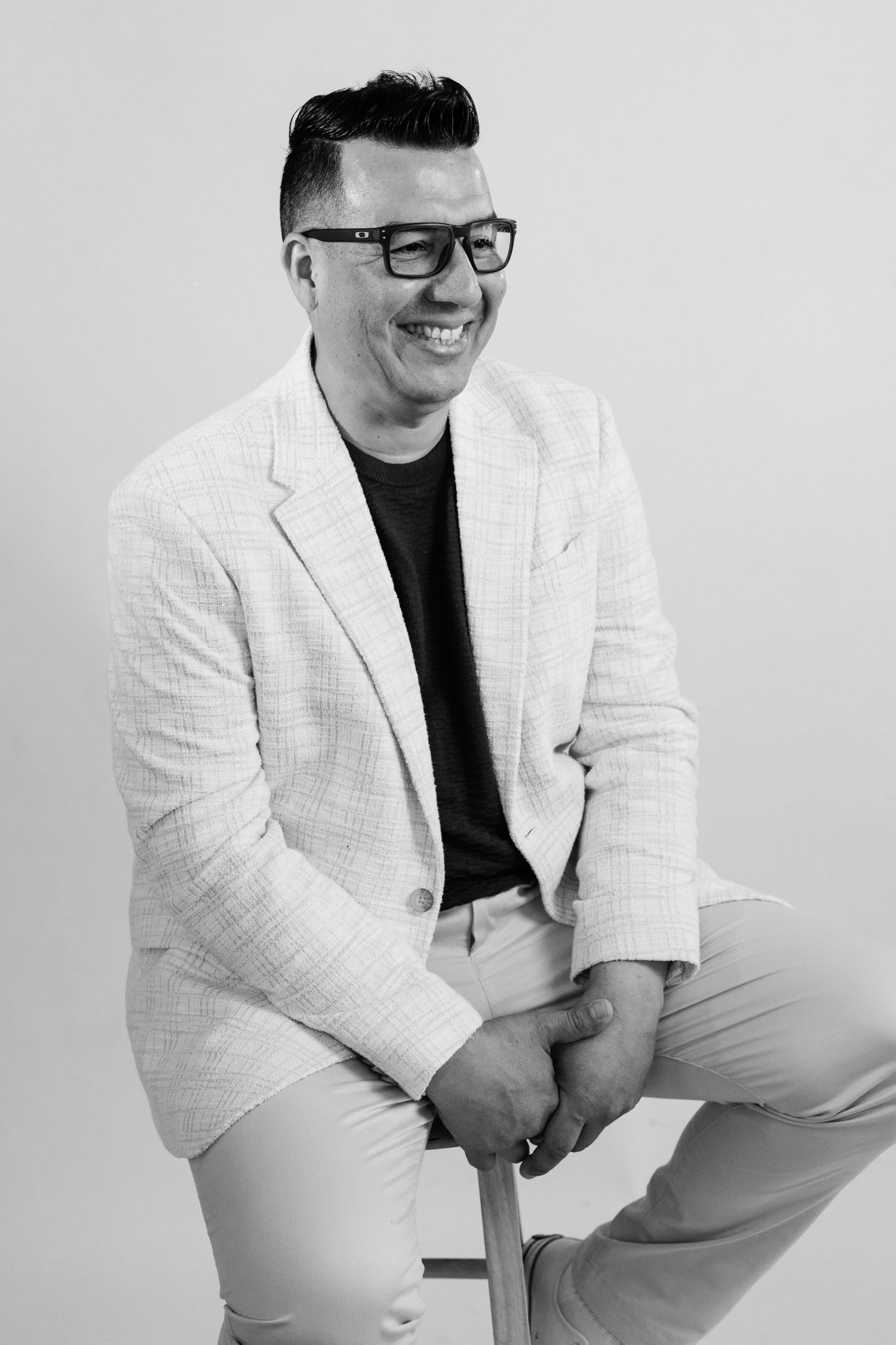 Black and white portrait of a smiling man in glasses and blazer sitting on a stool in a studio.