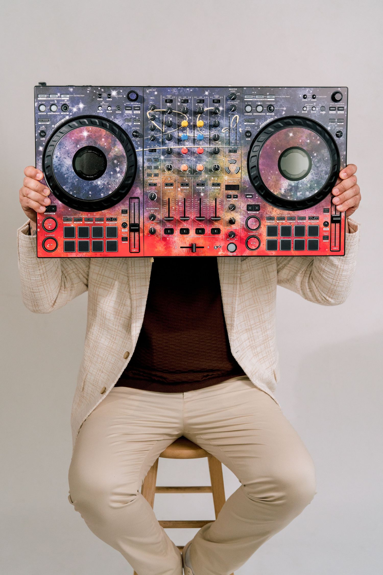 Person in beige suit sitting on stool holding colorful DJ mixer controller obscuring their face.