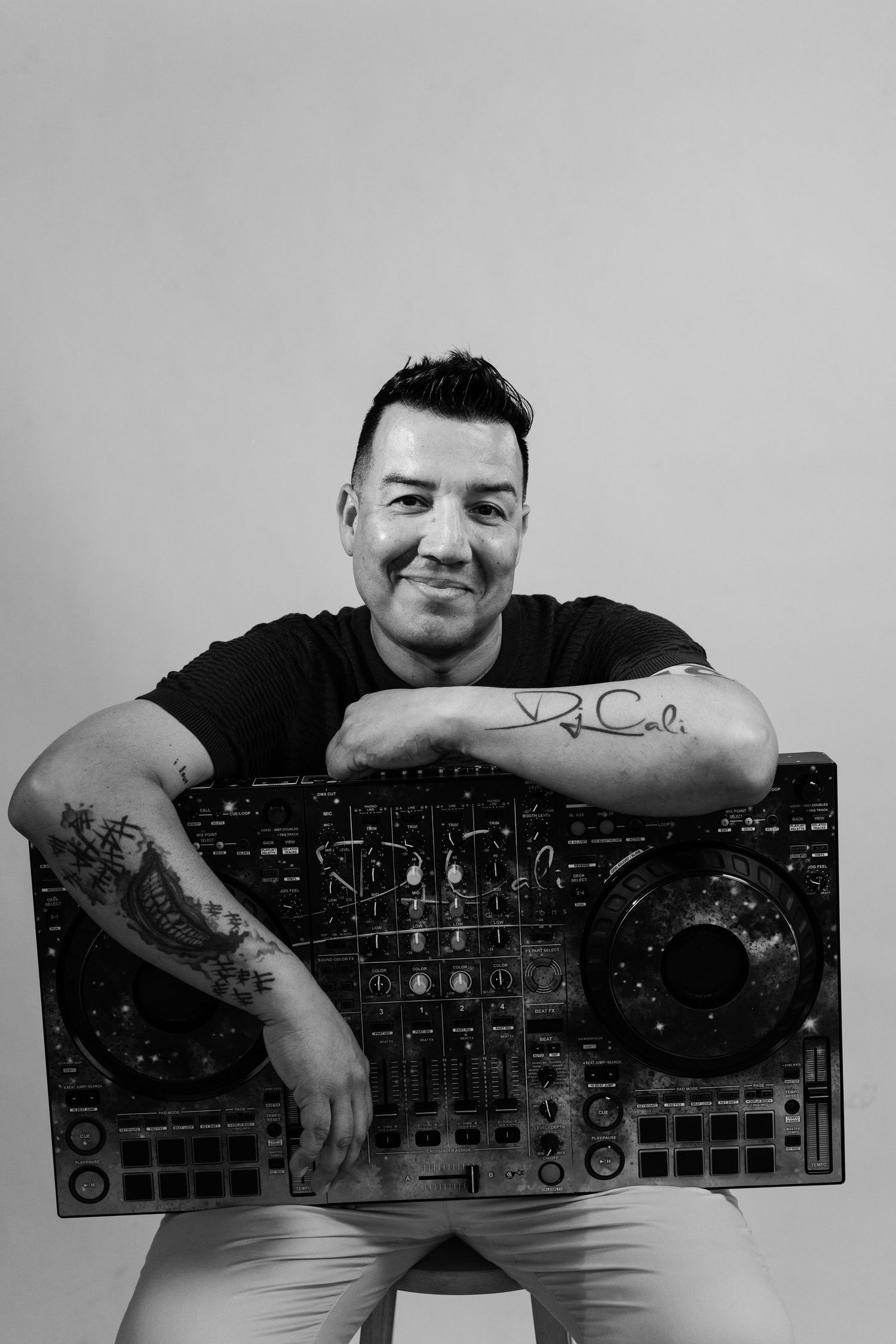 Black and white photo of a smiling male DJ posing with a Pioneer DJ controller, showing tattoos on both arms.