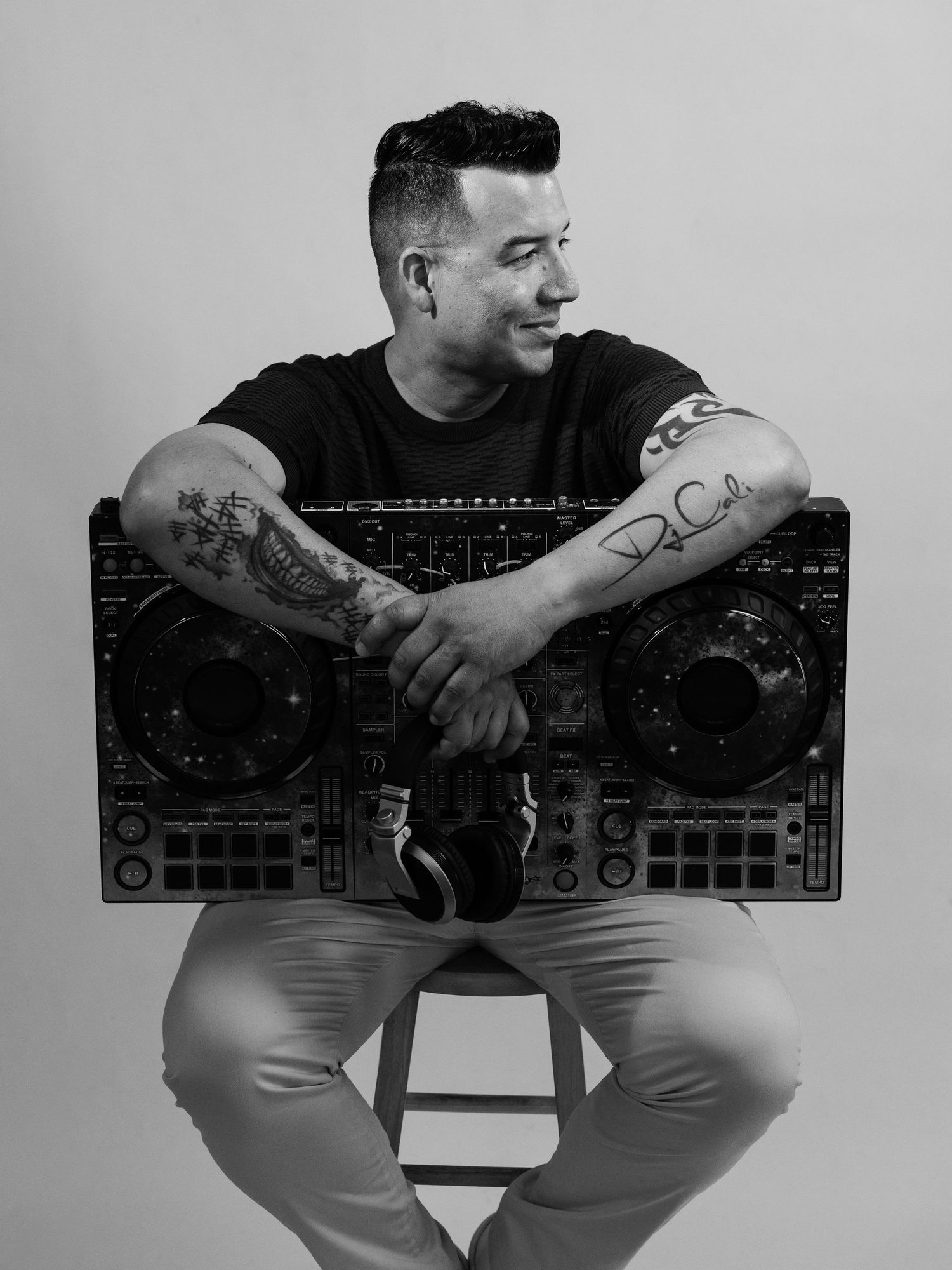 Black and white photo of a tattooed DJ posing with a Pioneer DJ controller while seated on a stool.