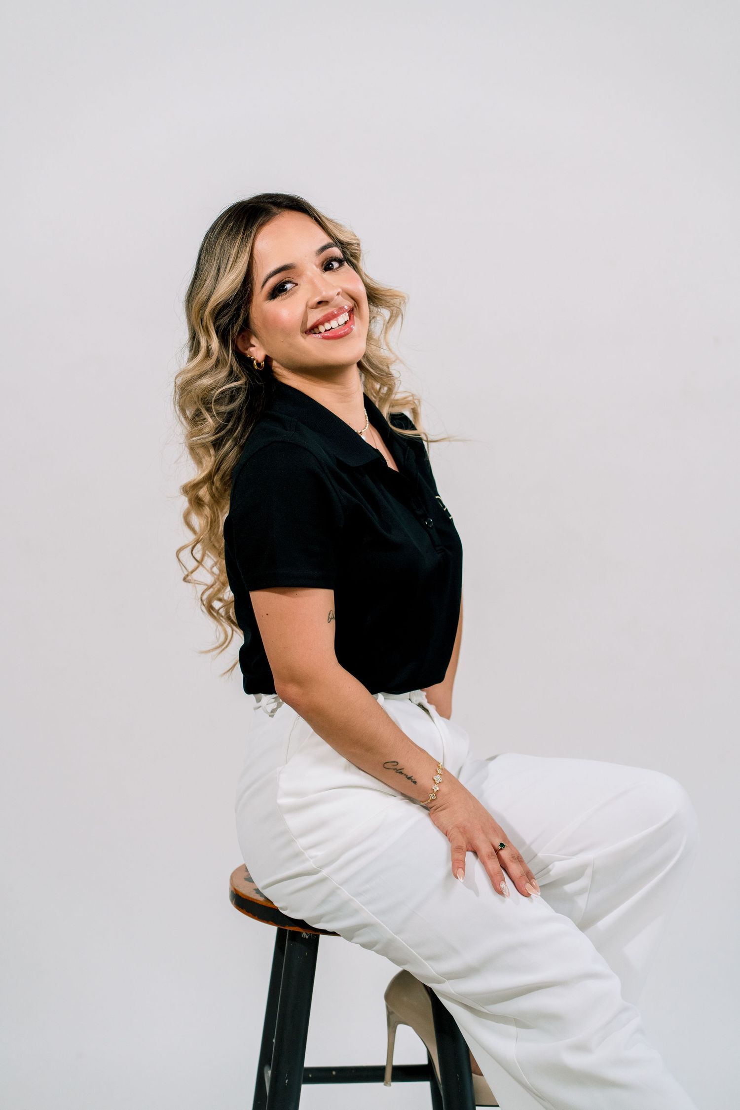 Smiling woman with curly blonde hair sitting on a stool, wearing black top and white pants against gray background.