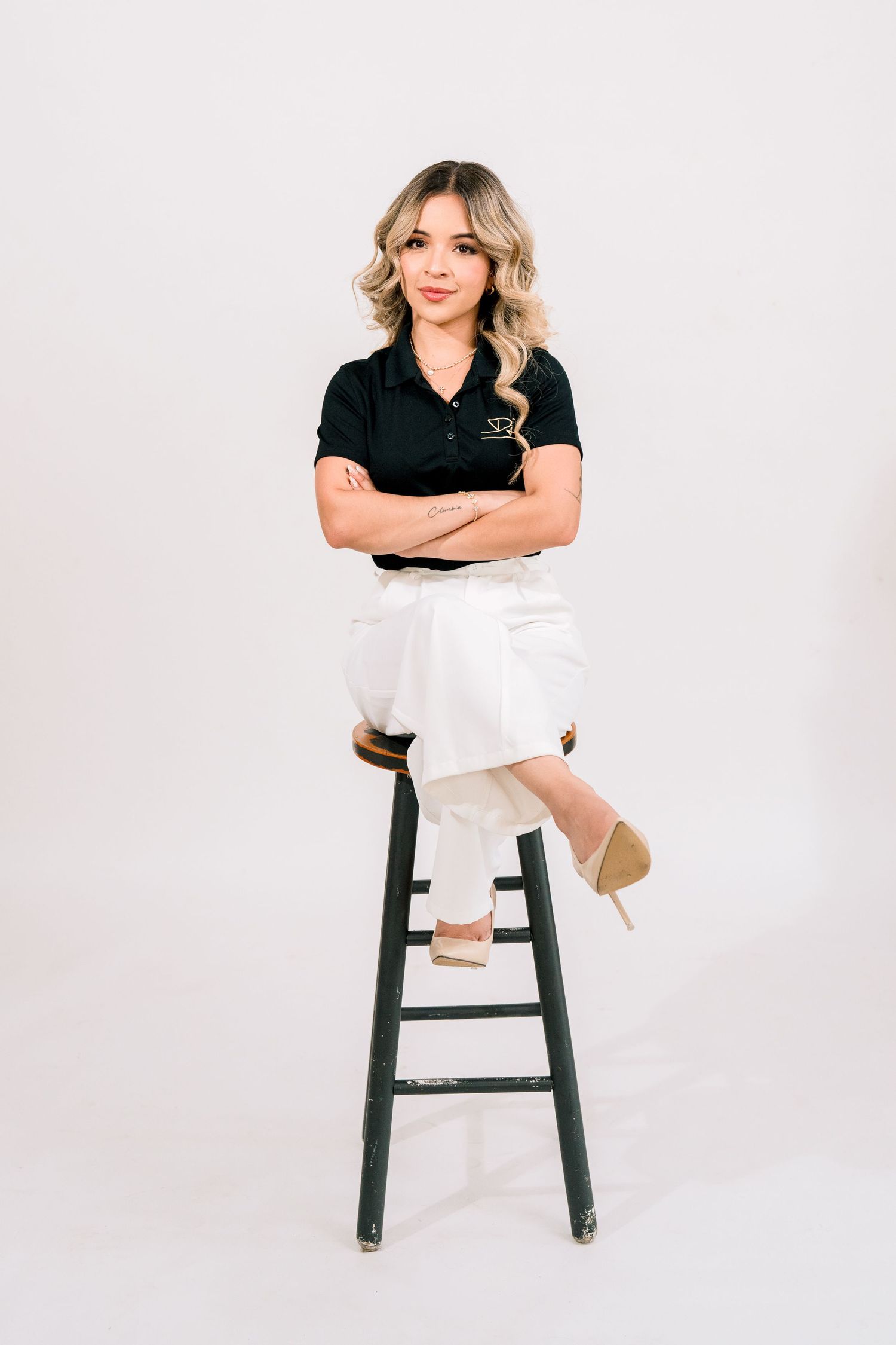 Woman in black polo and white pants sits cross-armed on a stool, posing confidently against a white background.