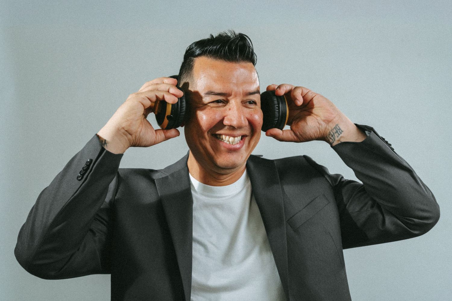 Smiling man in gray blazer holding black headphones to his ears against a light gray background.