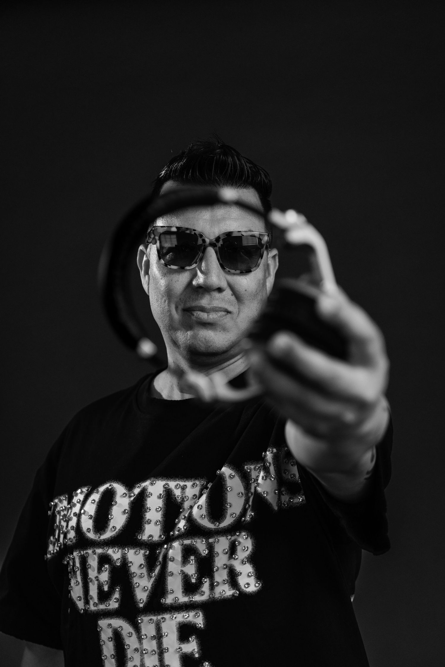 Black and white photo of a man in sunglasses and 'Emotions Never Die' shirt holding a camera toward viewer.