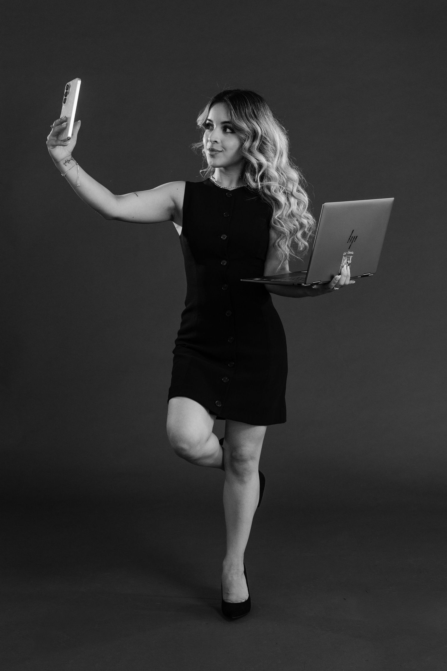 Young woman in black dress balancing on one leg, holding laptop and taking selfie with phone, black and white photo.