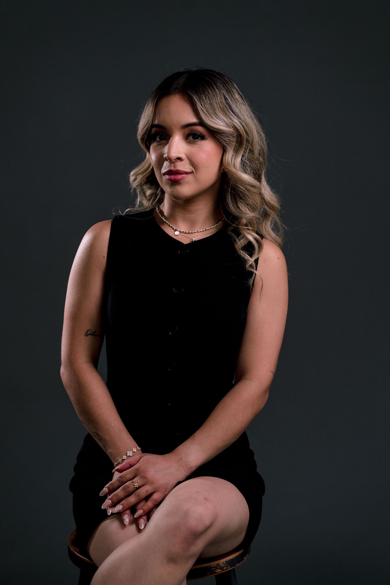 Young woman with blonde wavy hair in black sleeveless dress seated against dark gray studio background.
