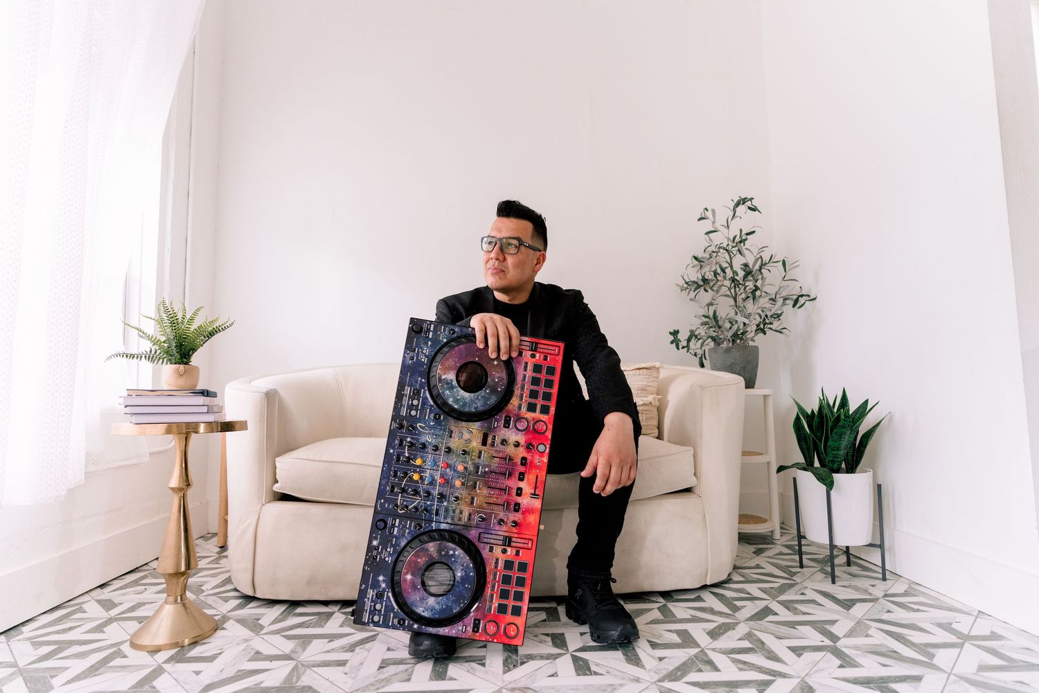 Man in black outfit sits on beige chair holding a colorful DJ controller in a bright modern living room.