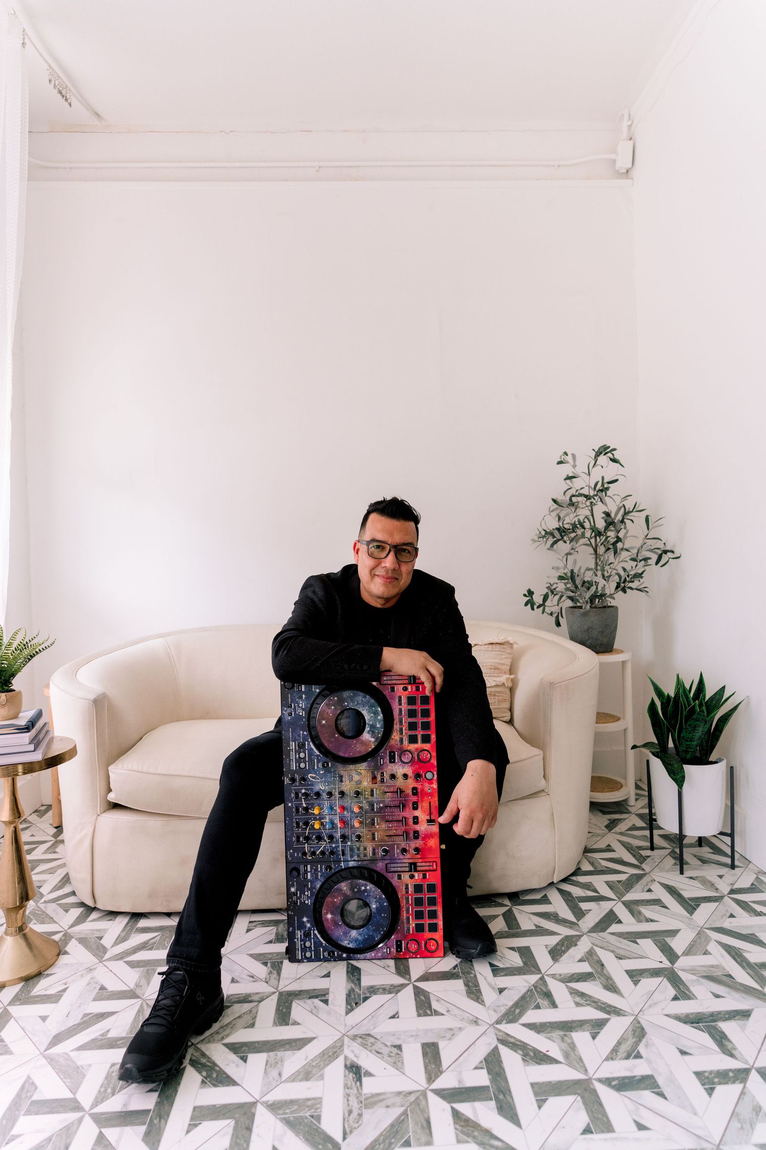 A DJ poses with a colorful mixer controller on patterned tile floor beside a cream sofa in a bright white room.