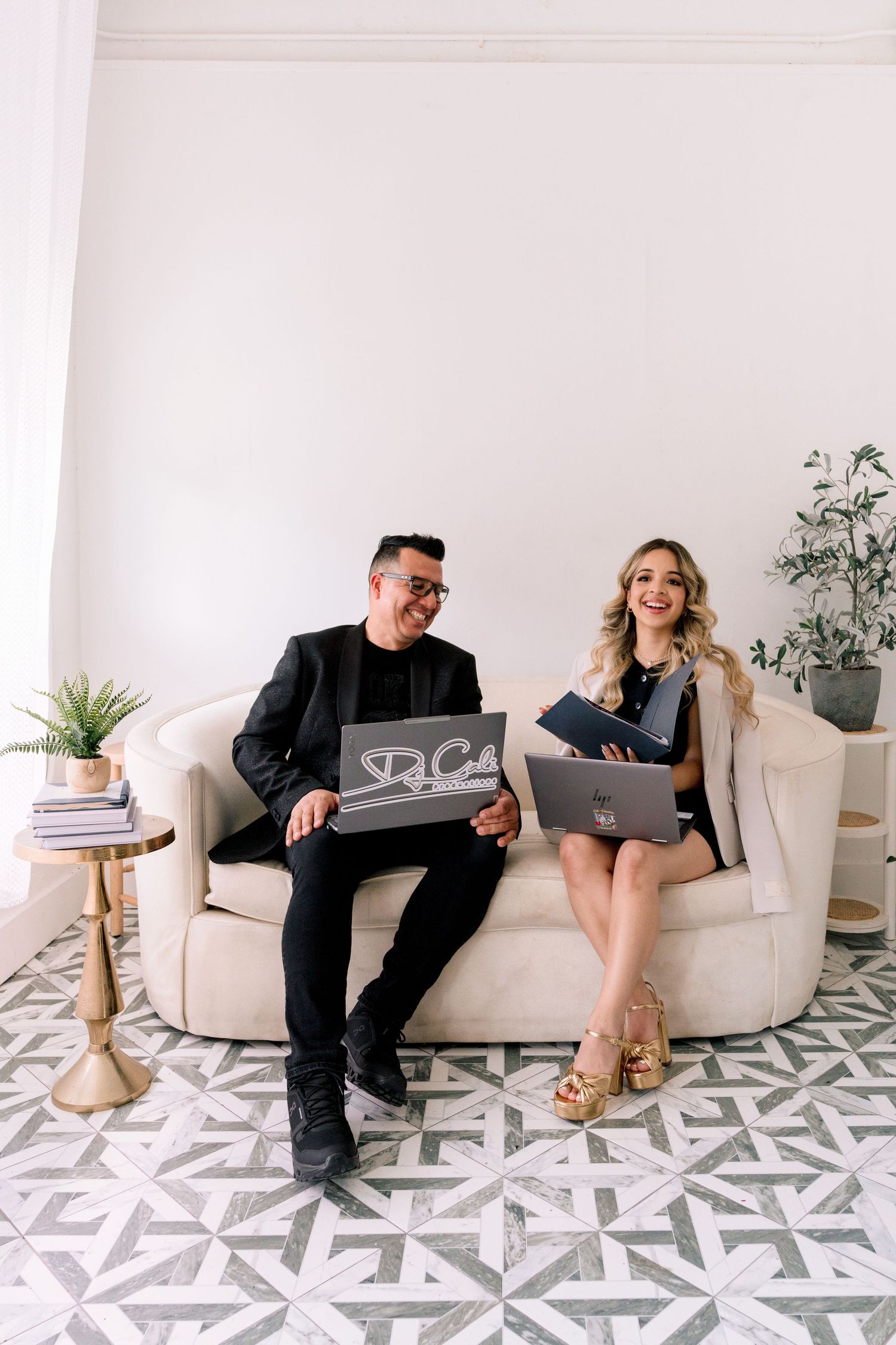 Man and woman sitting on elegant sofa holding a logo sign and laptop in a bright modern interior space.