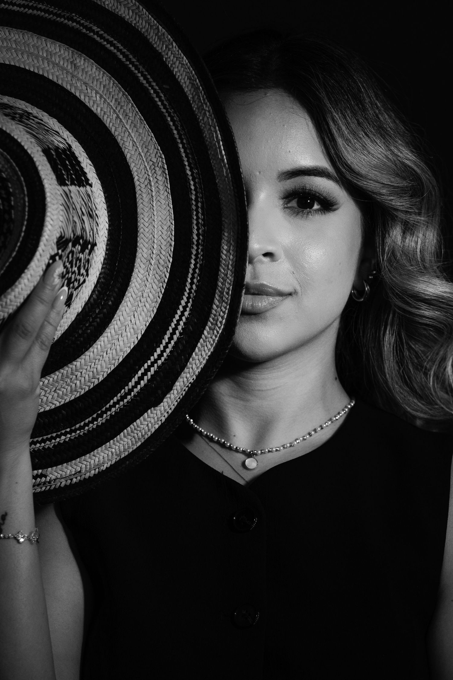 Black and white portrait of a woman holding a large striped sombrero hat beside her face in dramatic lighting.