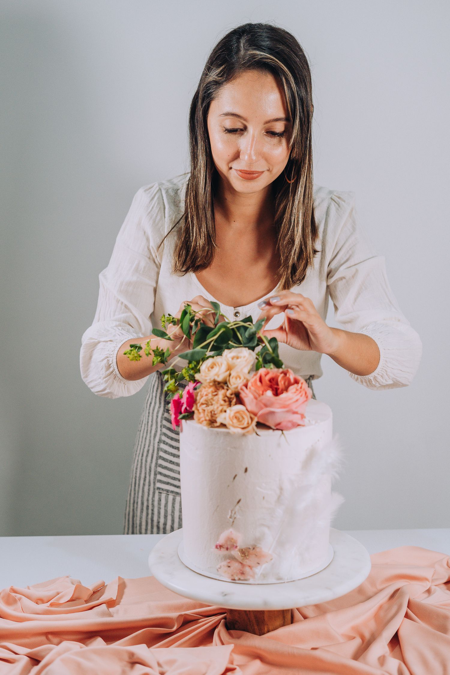 A cake decorator in a striped apron arranges fresh flowers on a white wedding cake with peach-colored fabric draping below.