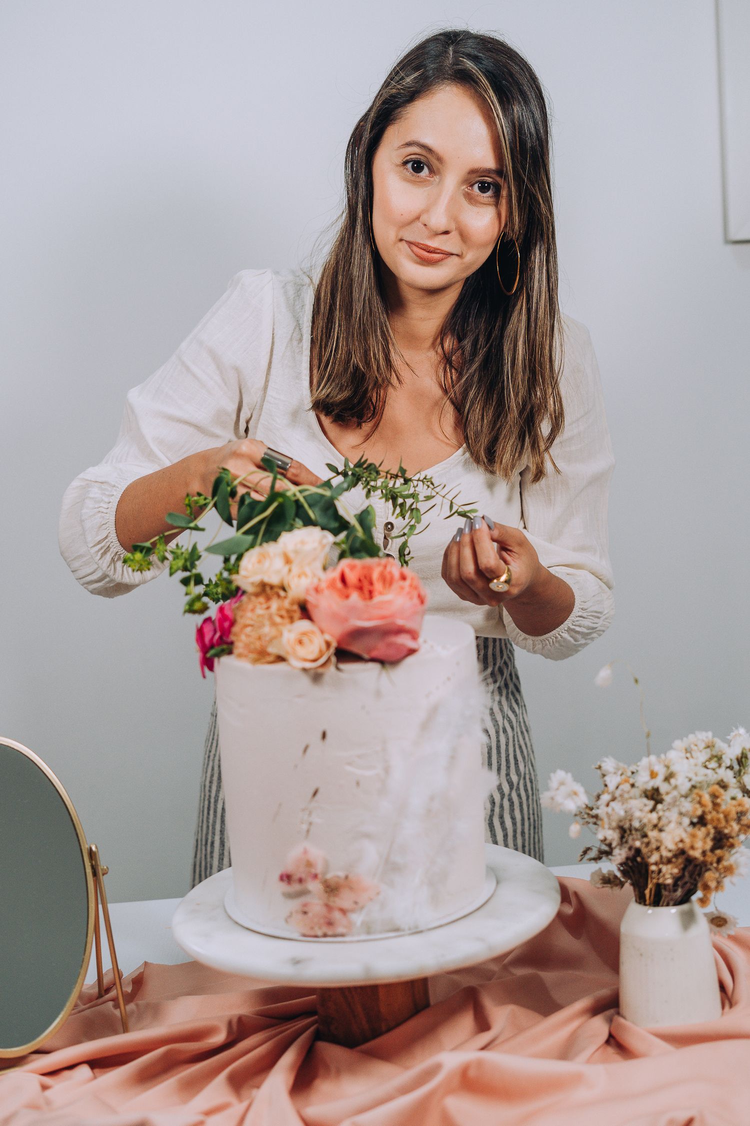 Someone decorates a white cake with fresh peach and pink flowers while wearing a white blouse and striped apron.