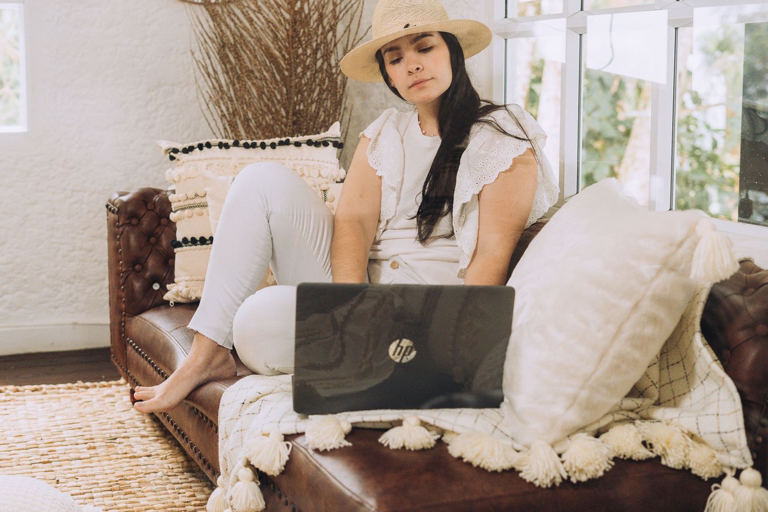 Cozy home office setup with laptop, brown leather couch, and decorative throw pillows in natural light.