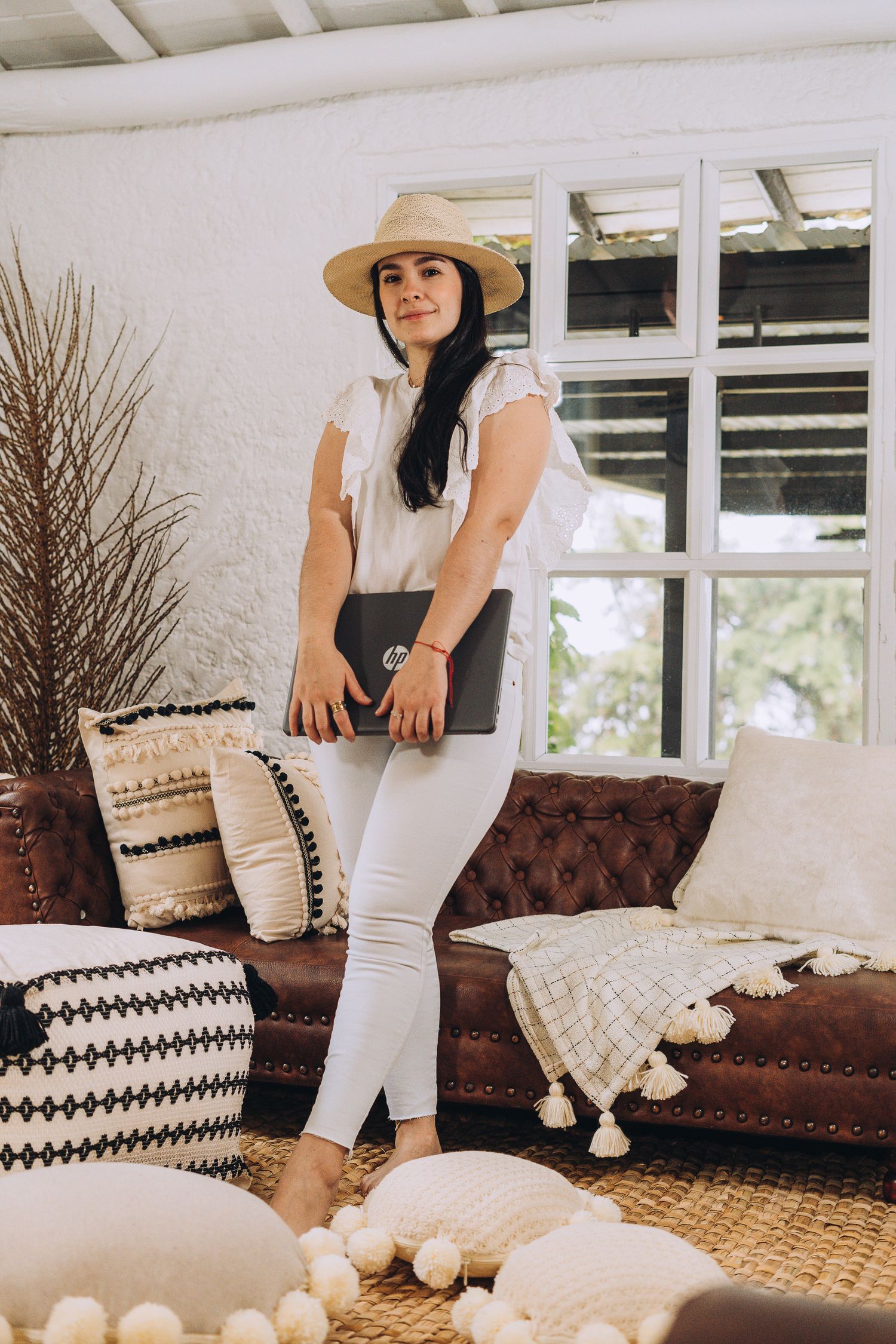 Lifestyle photo of a casual outfit with a straw hat, white blouse, and brown leather couch in a bright sunlit room.