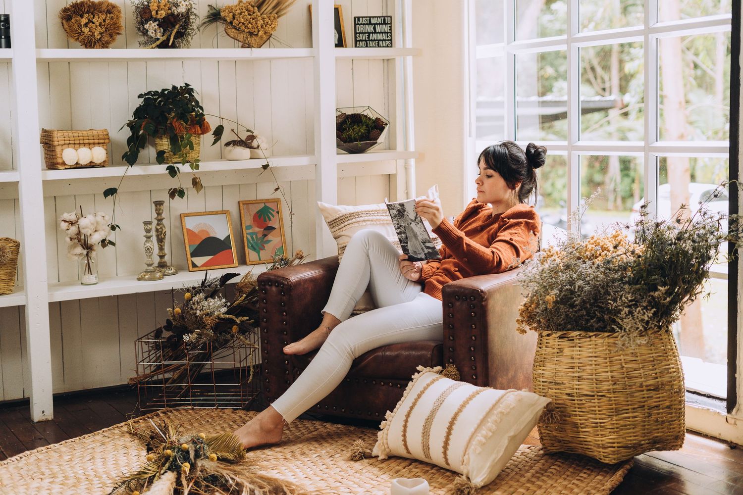A cozy reading nook with white shelves, wicker basket, and someone reading in a comfortable armchair by large windows.