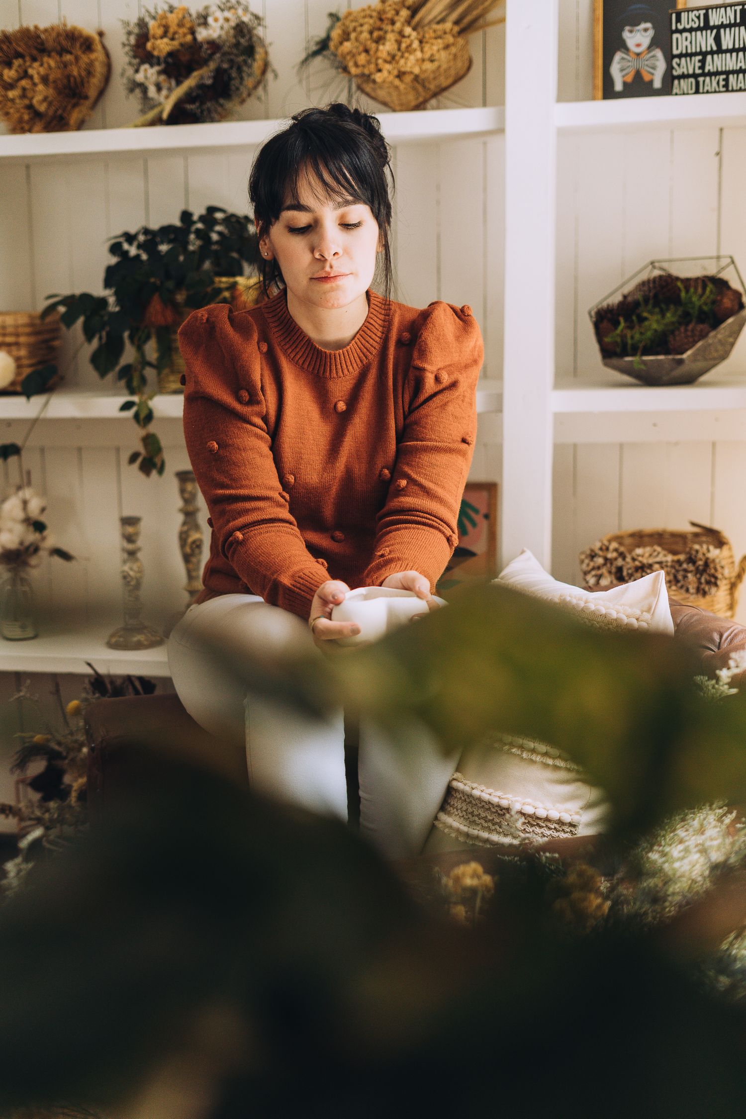 A person in a rust-colored sweater sits quietly in a room decorated with plants and shelving displays.