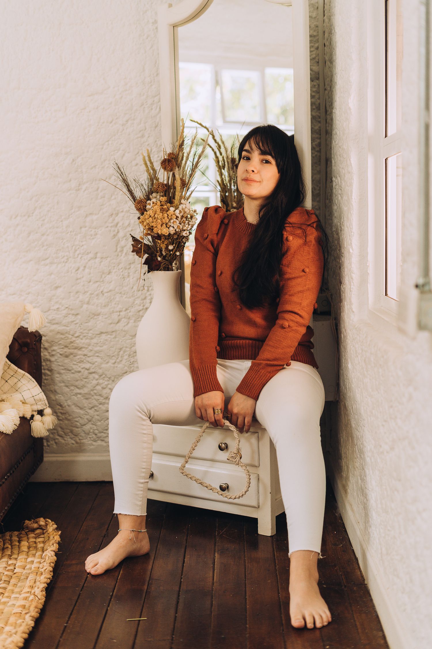 A person in a rust-colored sweater and white pants sits barefoot on a small white dresser near dried floral arrangements.