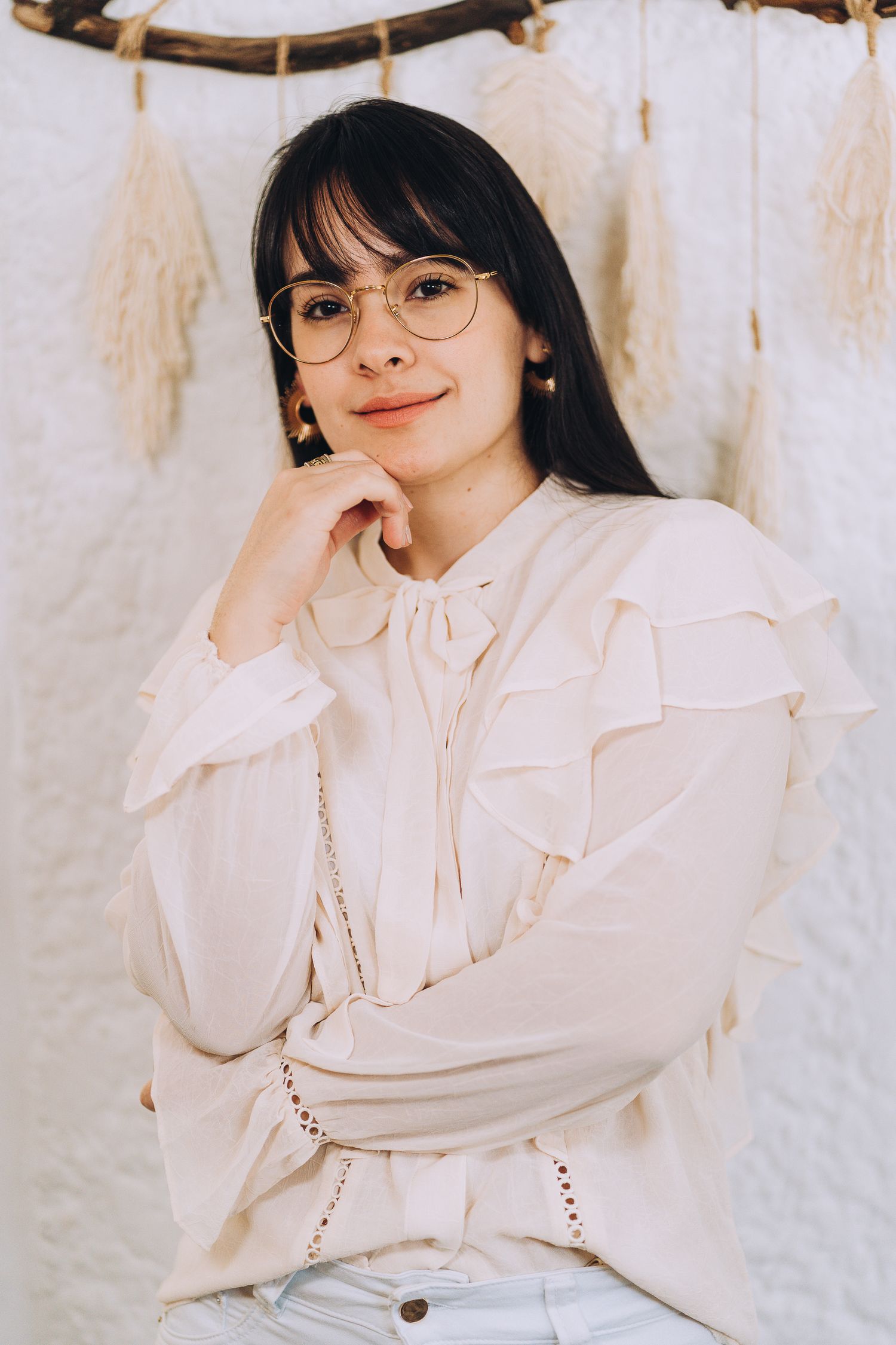 A person wearing glasses and a white ruffled blouse poses against a light backdrop with macramé wall decor.