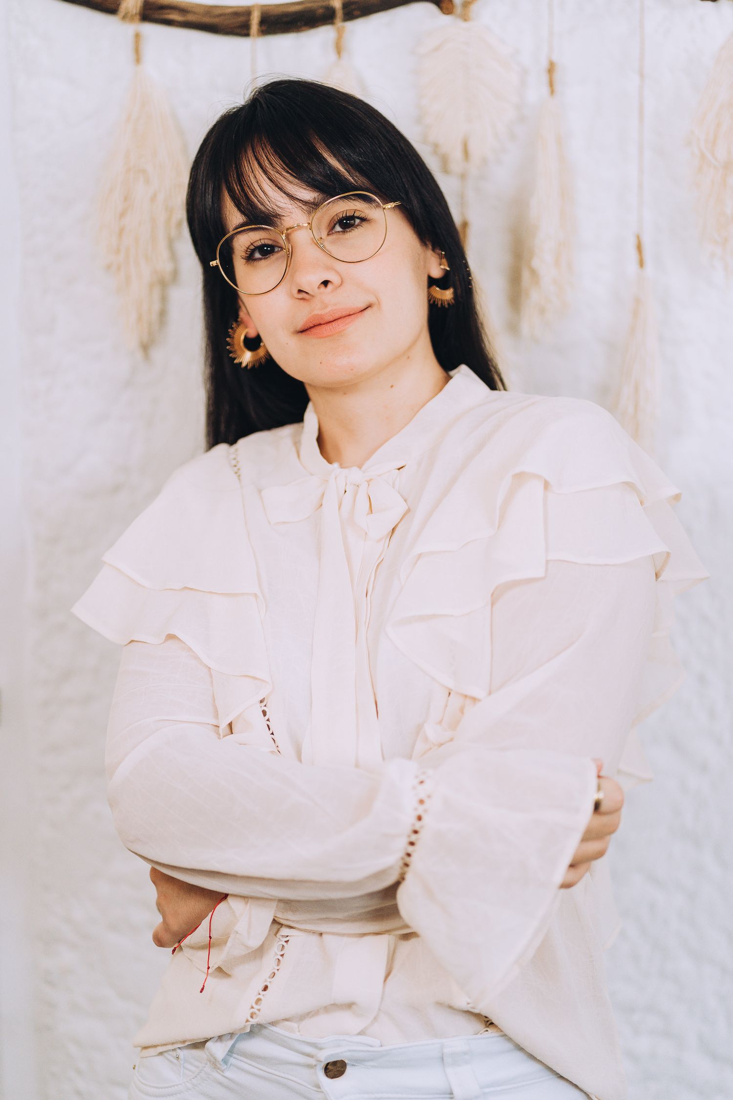 A person wearing a white ruffled blouse and round glasses poses against a light background.