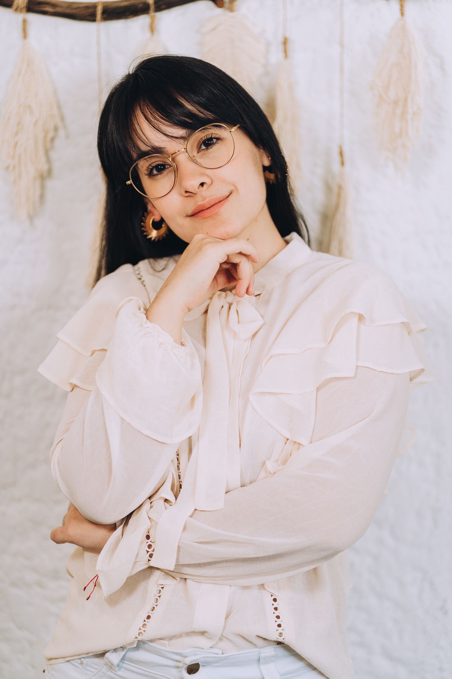 A portrait in soft lighting features someone wearing a white ruffled blouse and round glasses against a pale background.