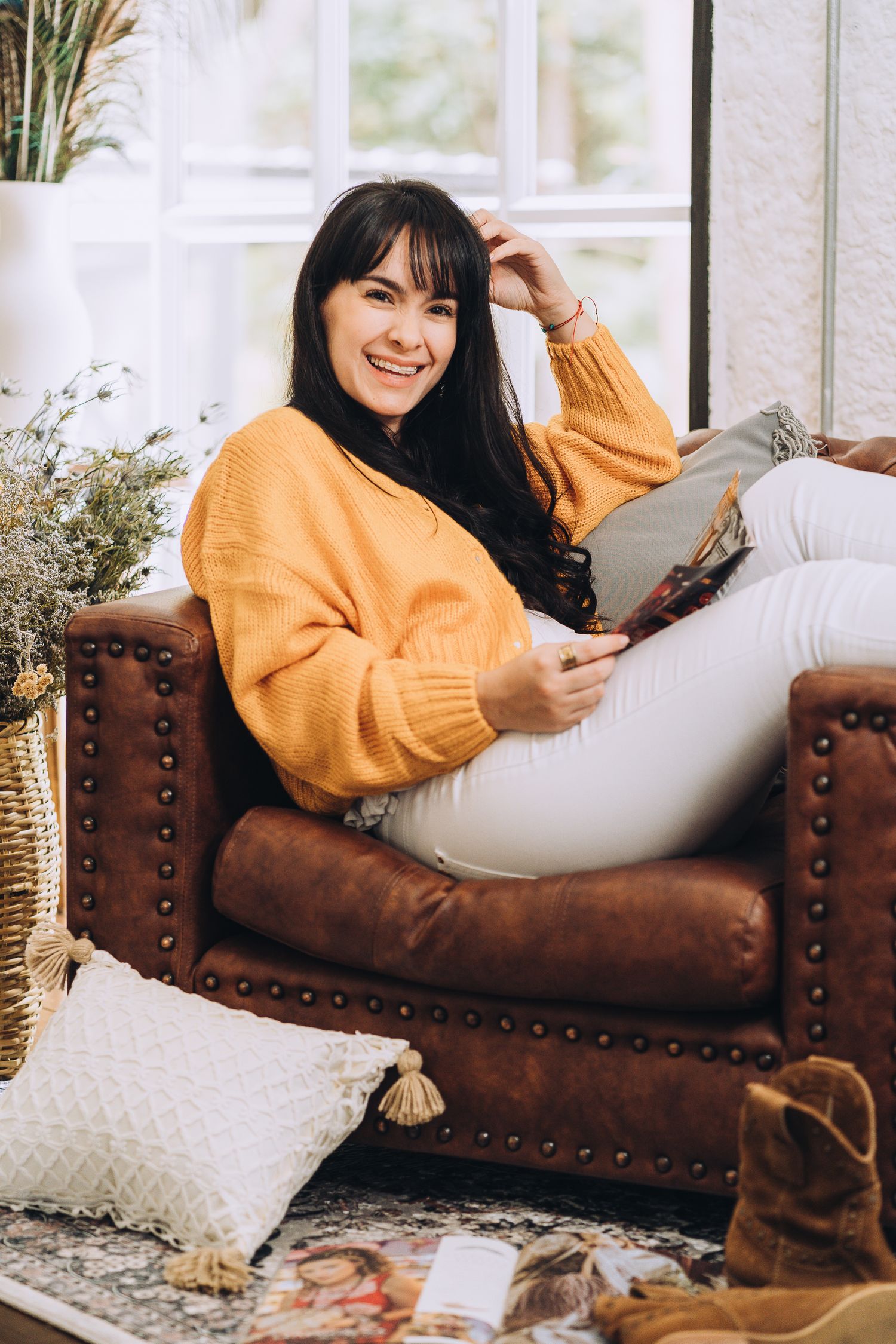 A person in a cozy yellow sweater relaxes with a book on a brown leather chair by a window.