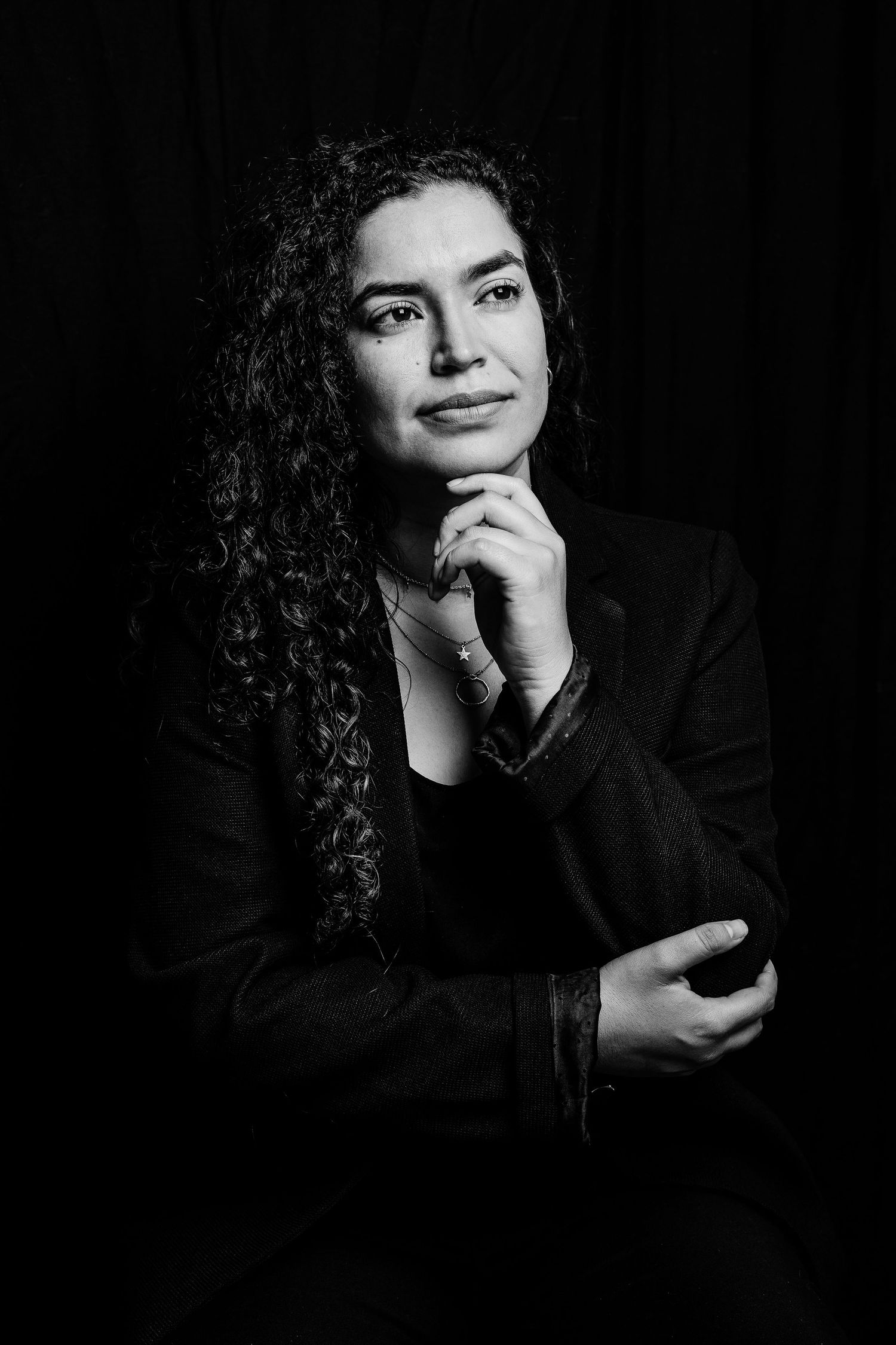 Professional portrait of a conductor in a black blazer with curly hair against dark background in contemplative pose.
