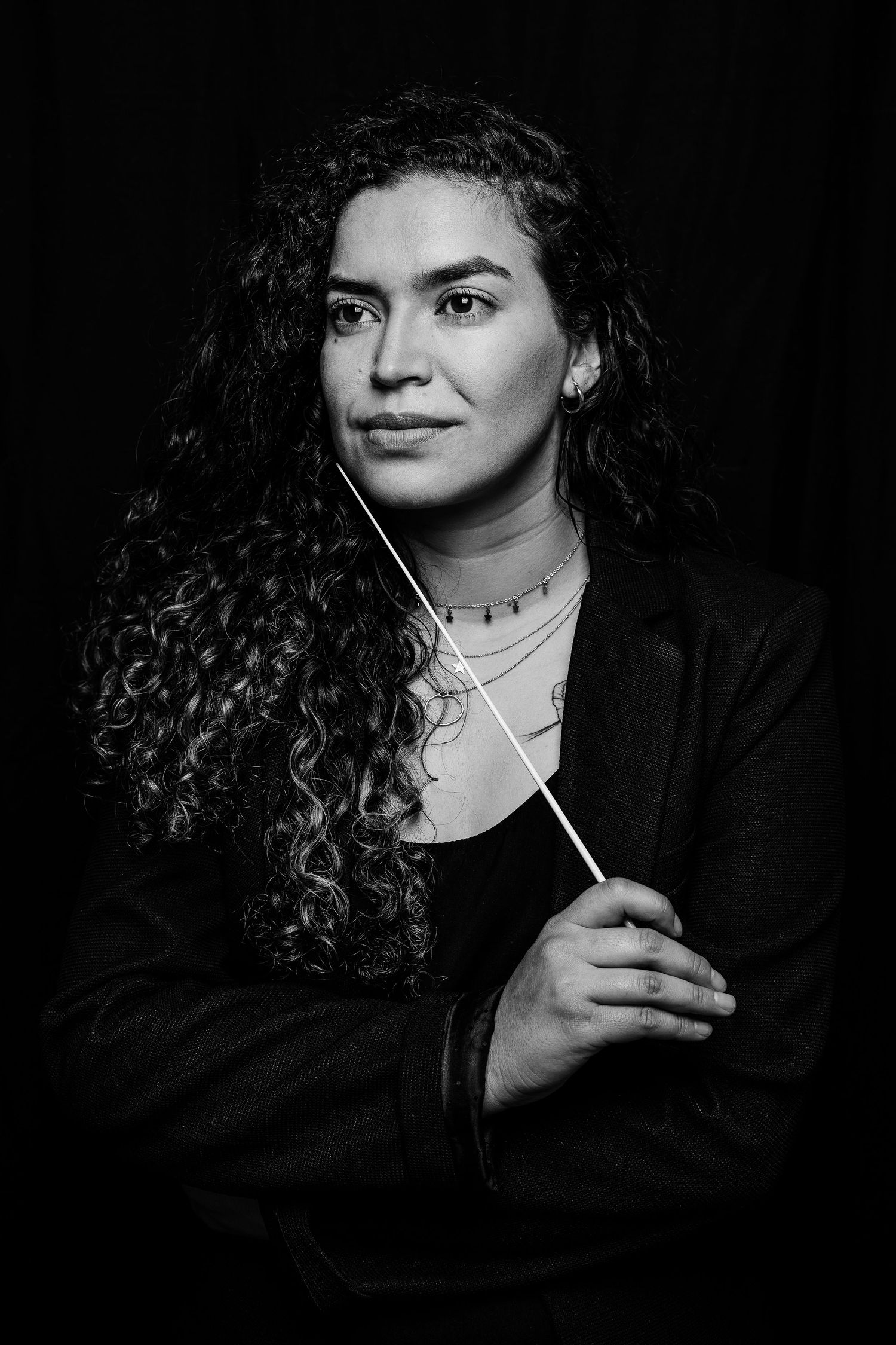 Conductor with long curly hair poses with baton in dramatic black and white portrait.