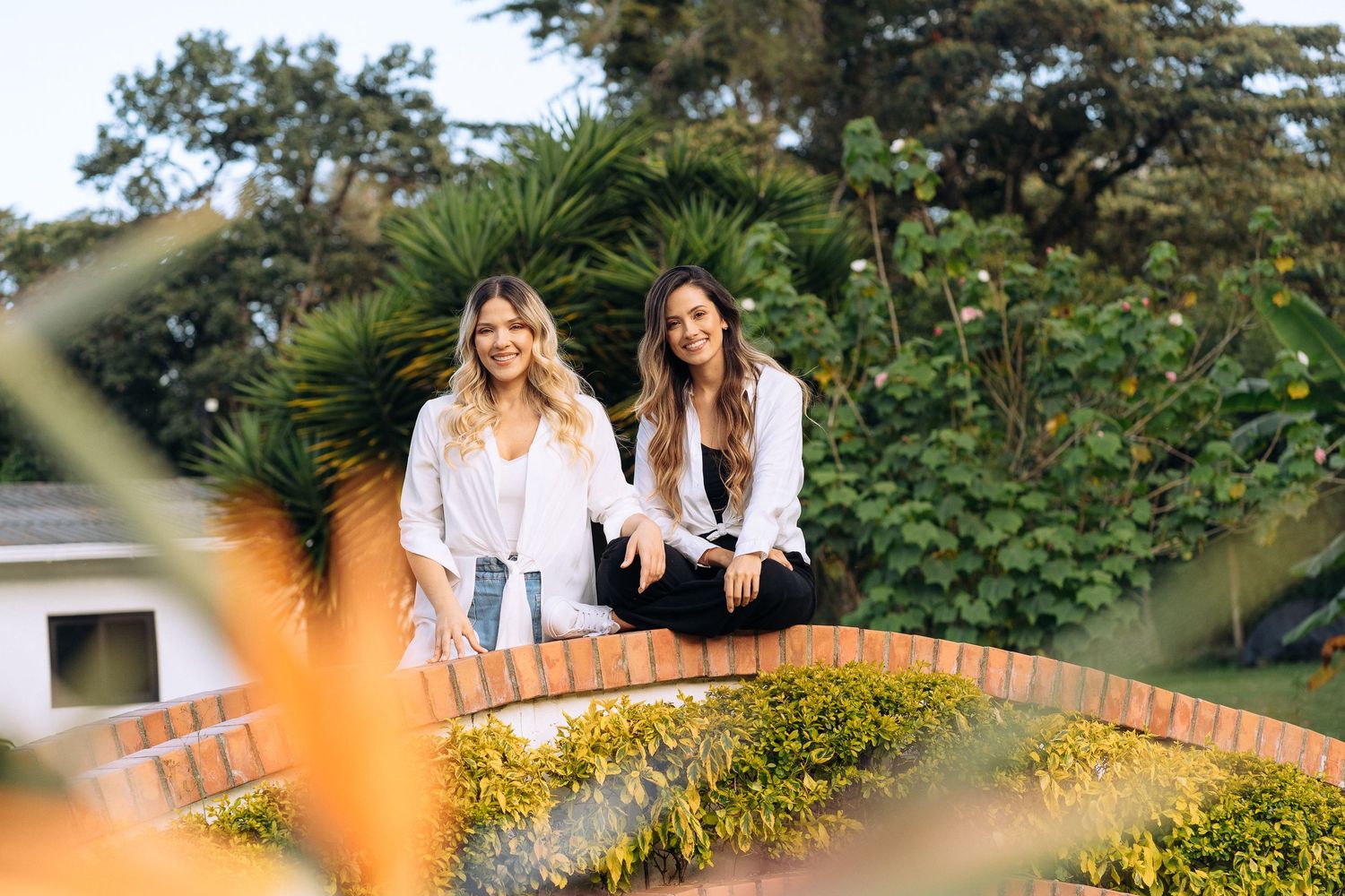 Two friends sitting on a decorative brick wall with tropical plants in background.