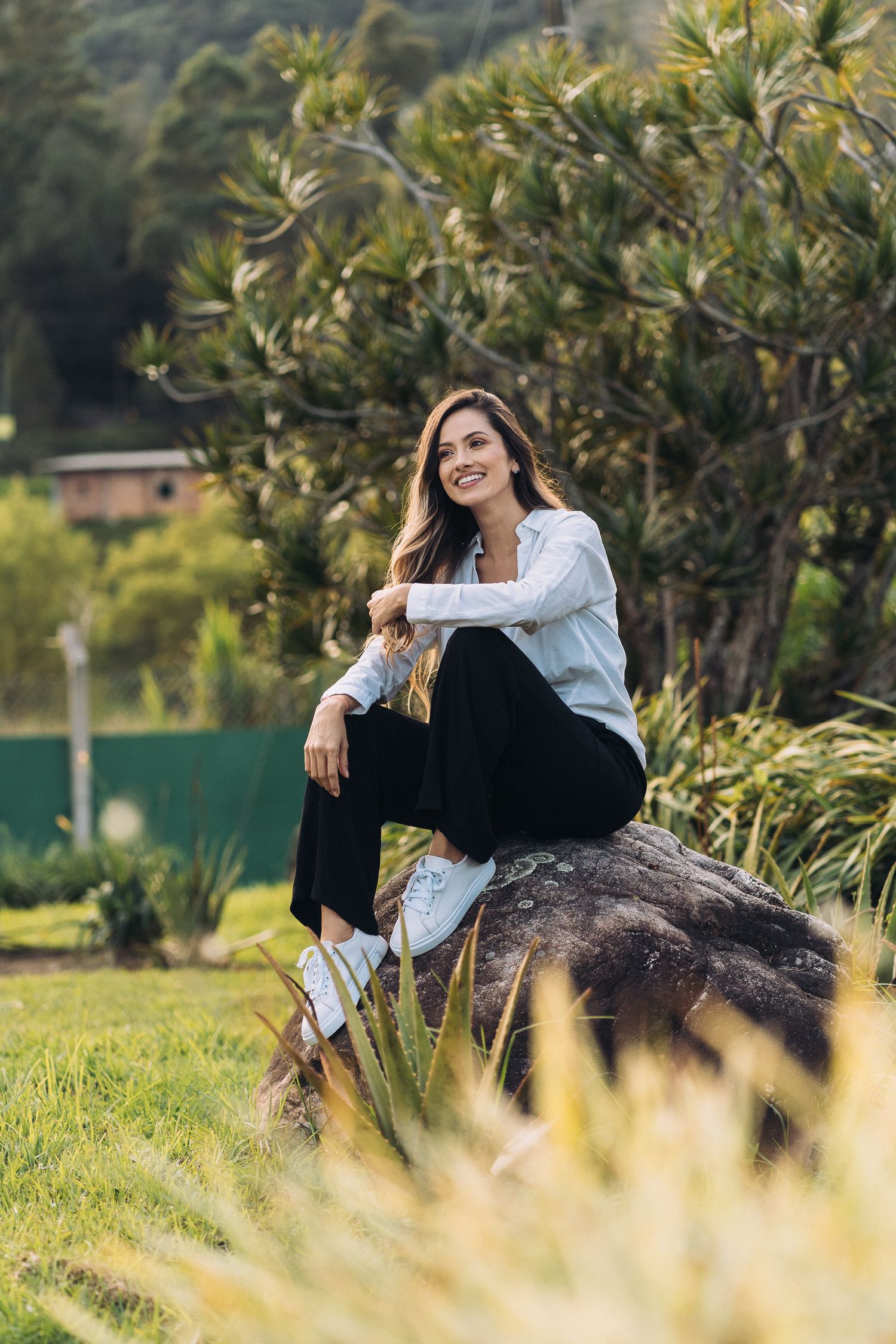 A person in a white shirt and black pants sits casually on a rock outdoors with lush greenery in the background.