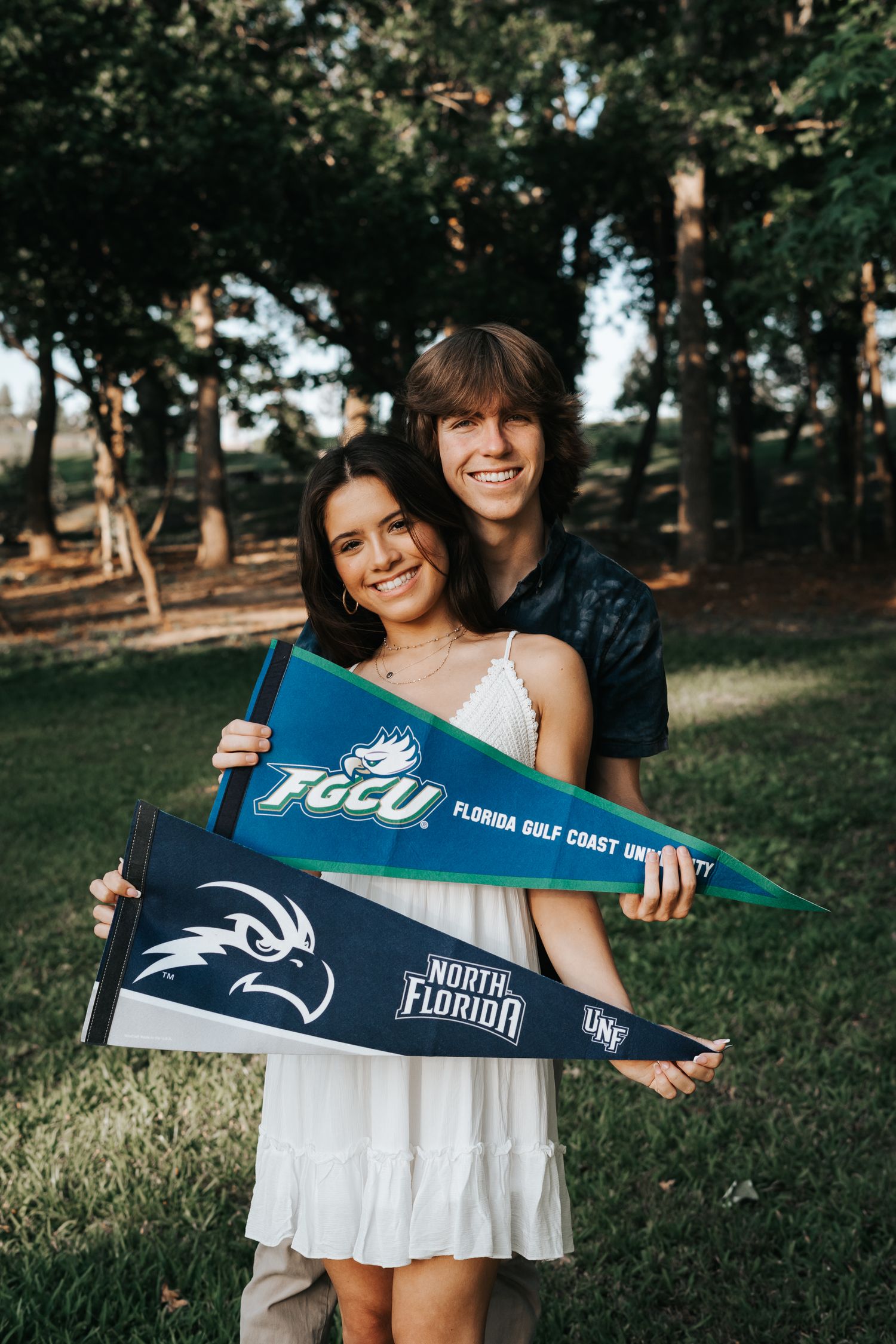 Couple showing school pride by holding FGCU Florida Gulf Coast University pennants outdoors in a wooded setting.