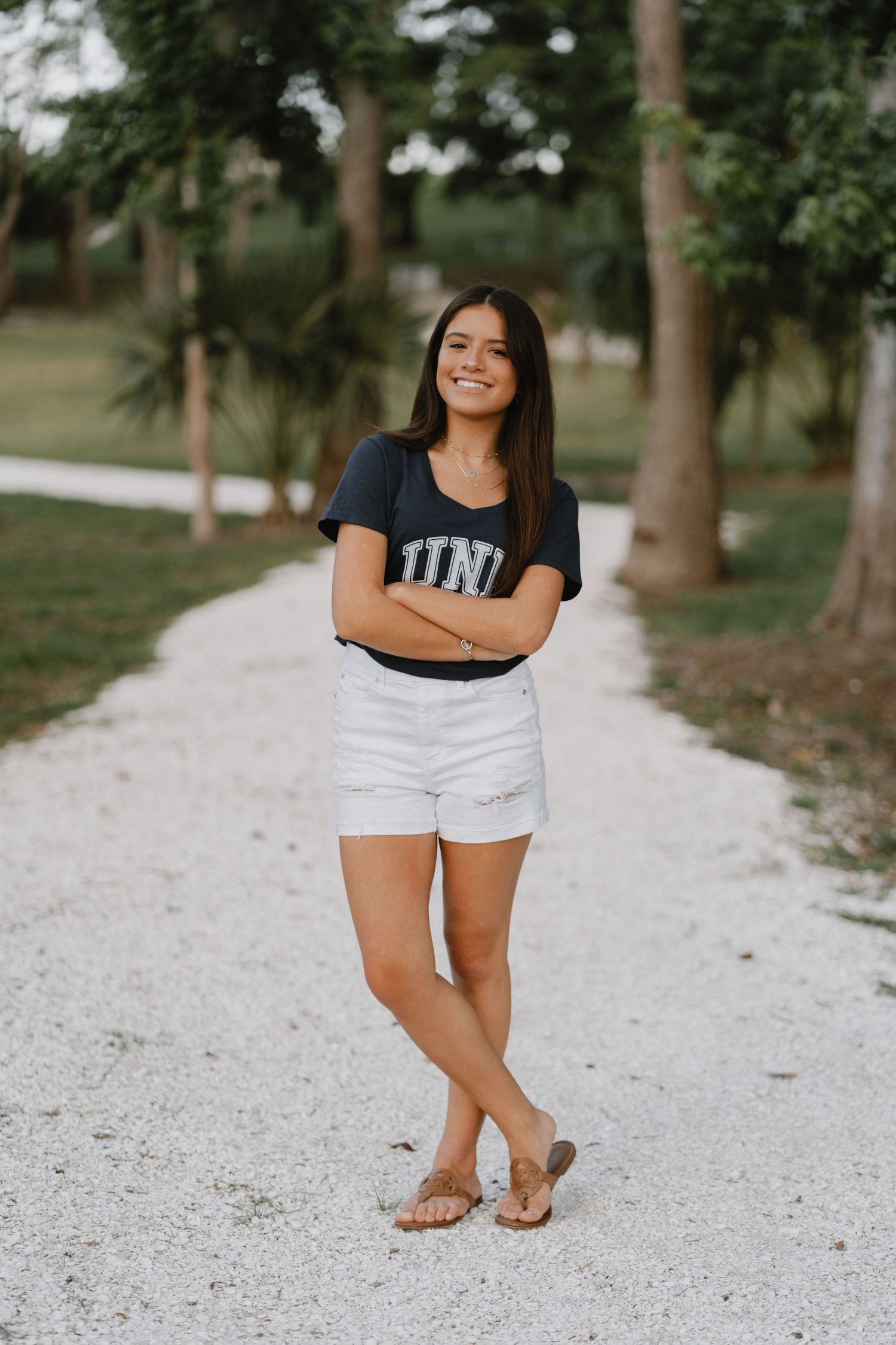 Student wearing UNF t-shirt and white shorts poses casually on a gravel path surrounded by trees.
