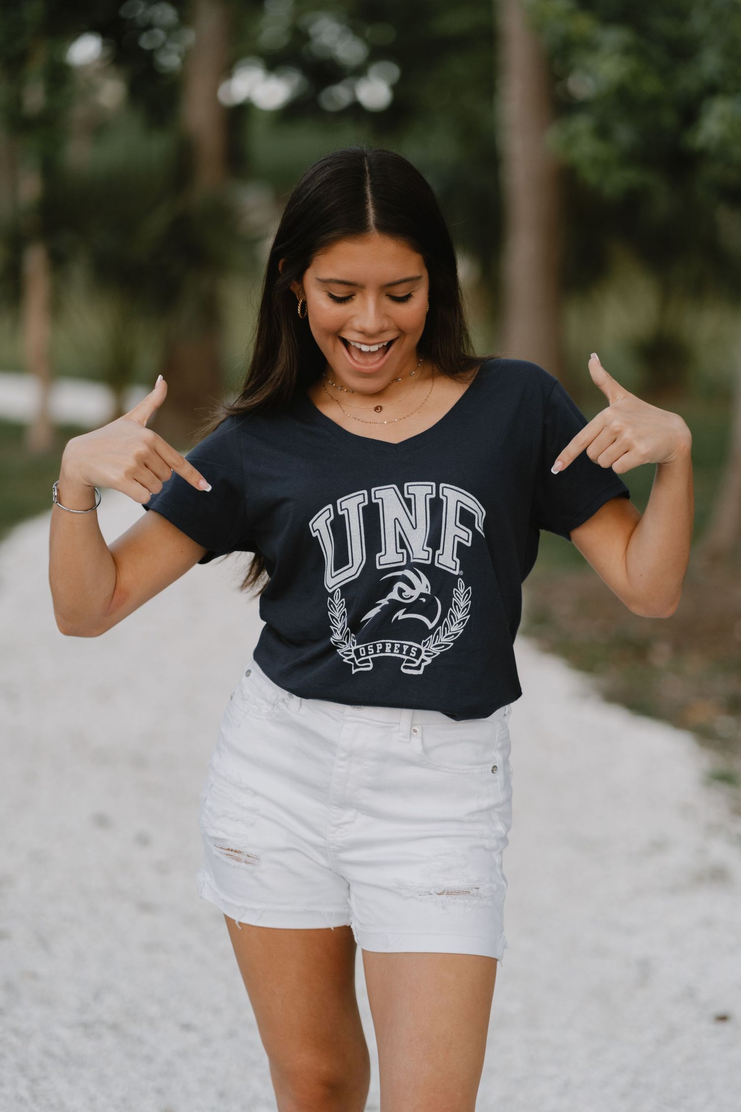 A person wearing a navy UNF athletic t-shirt and white distressed shorts poses pointing at the university logo outdoors.
