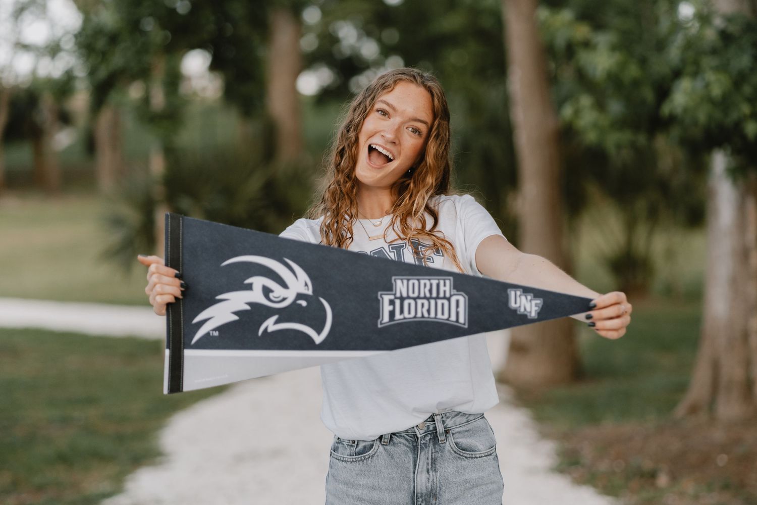An enthusiastic student displays a dark college pennant flag while standing outdoors on a sunny day.
