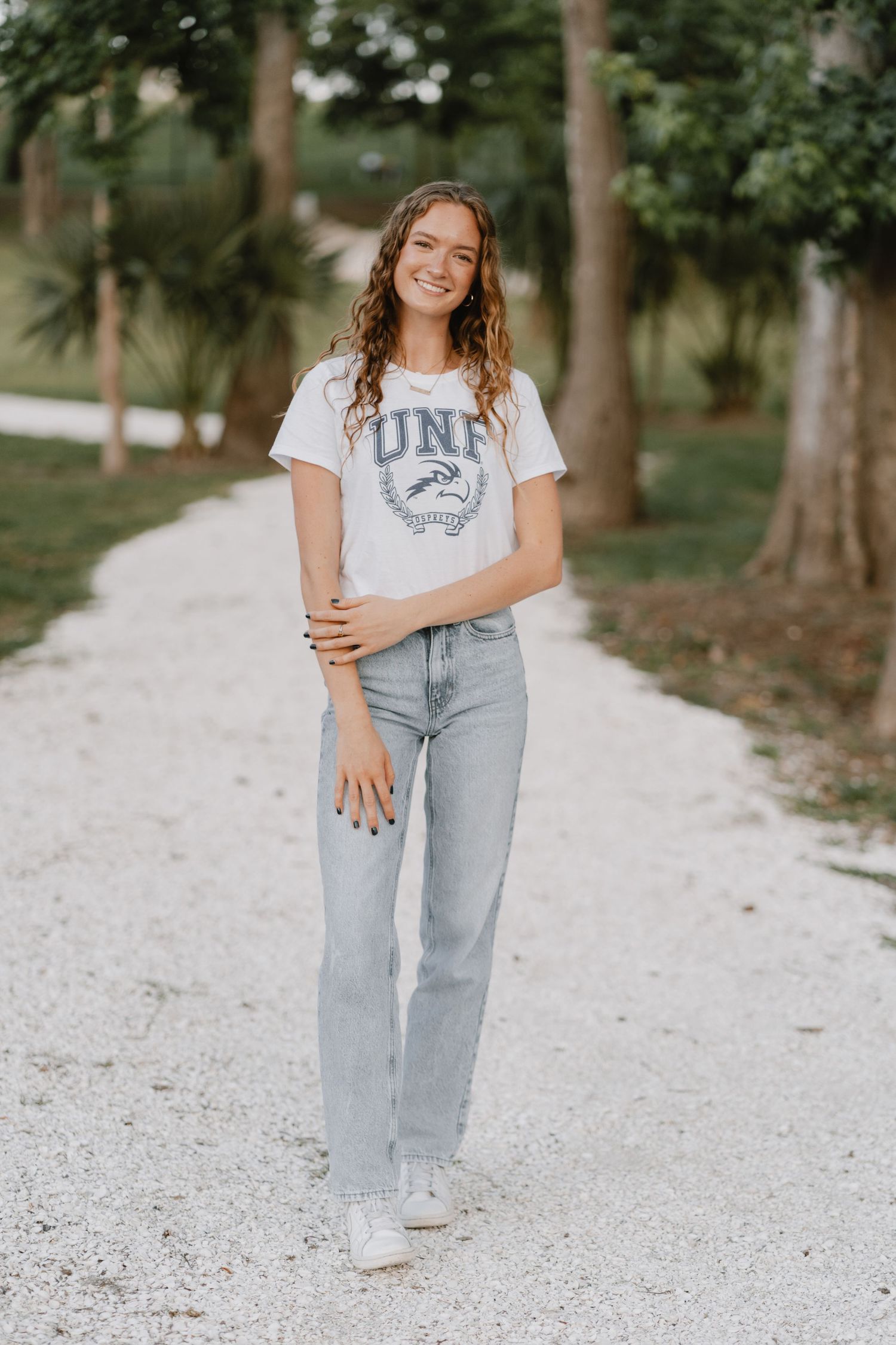 A person in a UNF t-shirt and light wash jeans stands on a white gravel path surrounded by trees.