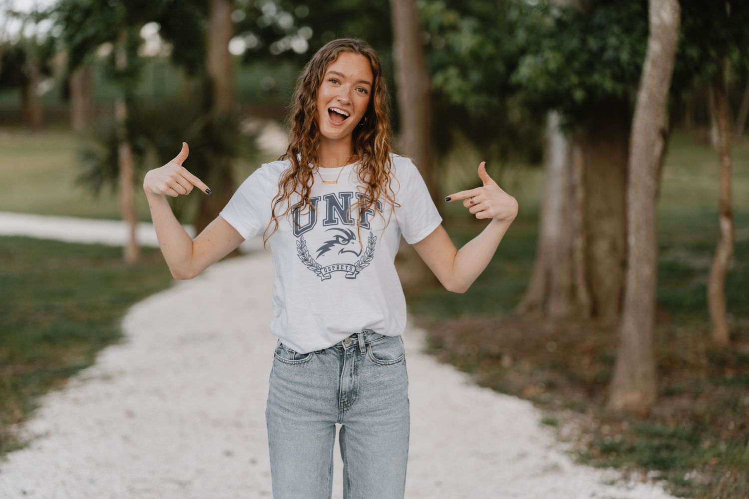 A playful person in light gray UNK t-shirt and jeans poses with thumbs up on a gravel path surrounded by trees.