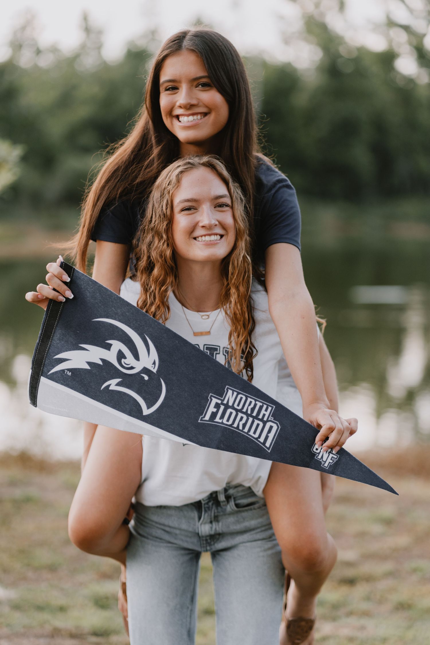 Two students smile while holding a North Florida college pennant outdoors during a casual photo session.