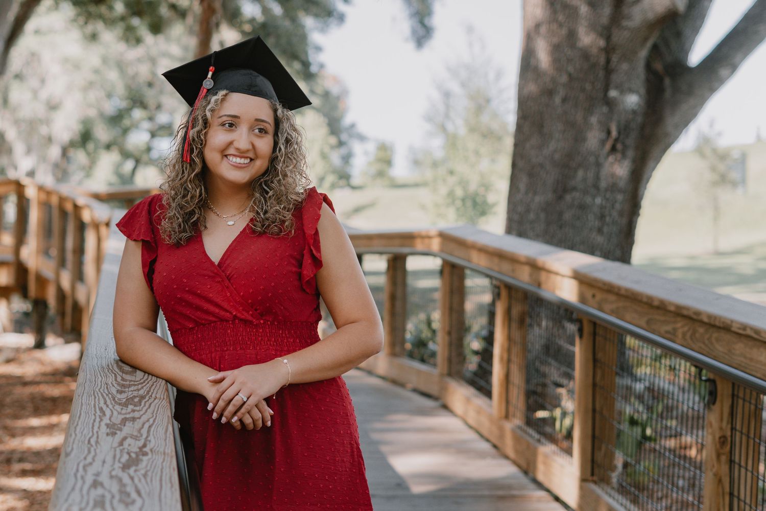A graduate wearing a red dress and graduation cap poses on a wooden deck surrounded by trees on a sunny day.
