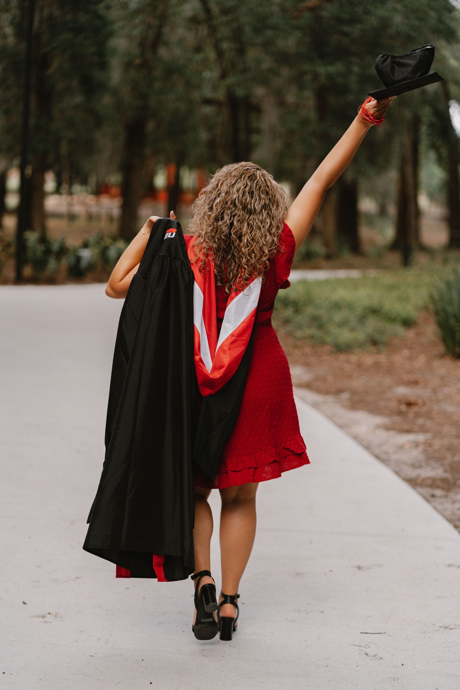 A graduate in a red dress walks down a path holding their graduation gown and cap triumphantly in the air.
