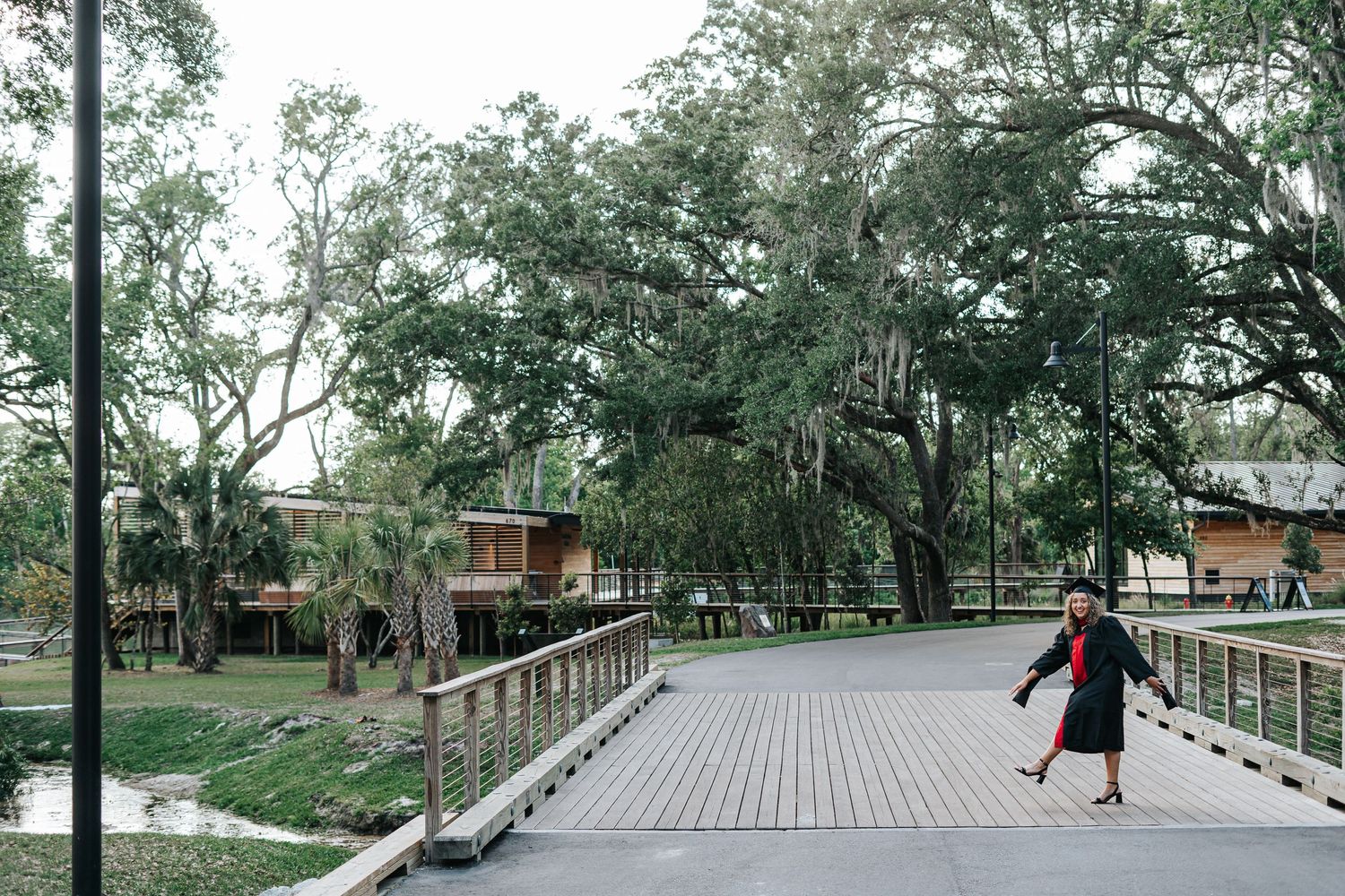A person skateboards on a wooden boardwalk path surrounded by Spanish moss-draped trees in a lush park setting.