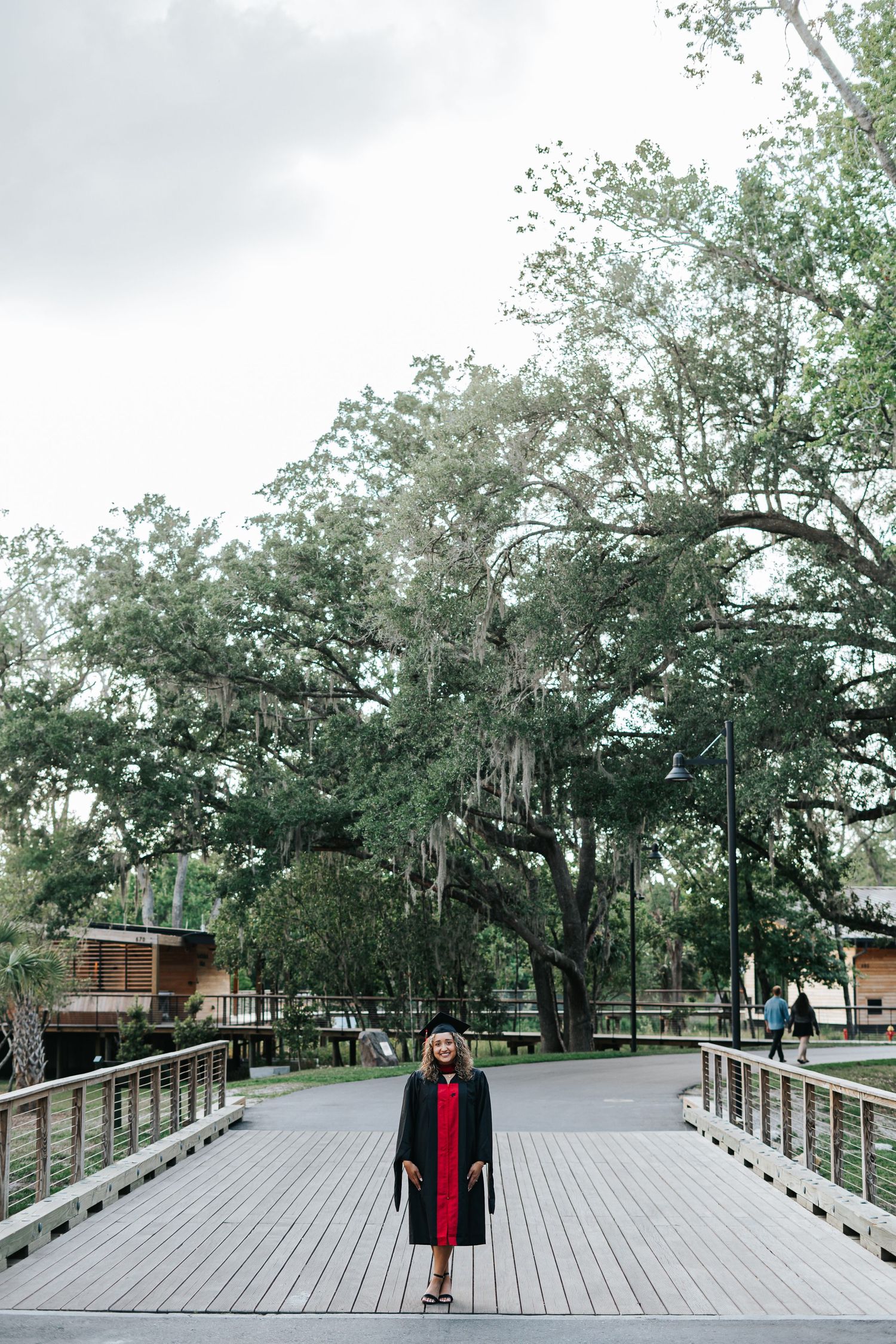 A person in a red coat stands on a wooden boardwalk surrounded by moss-draped oak trees at a nature park.