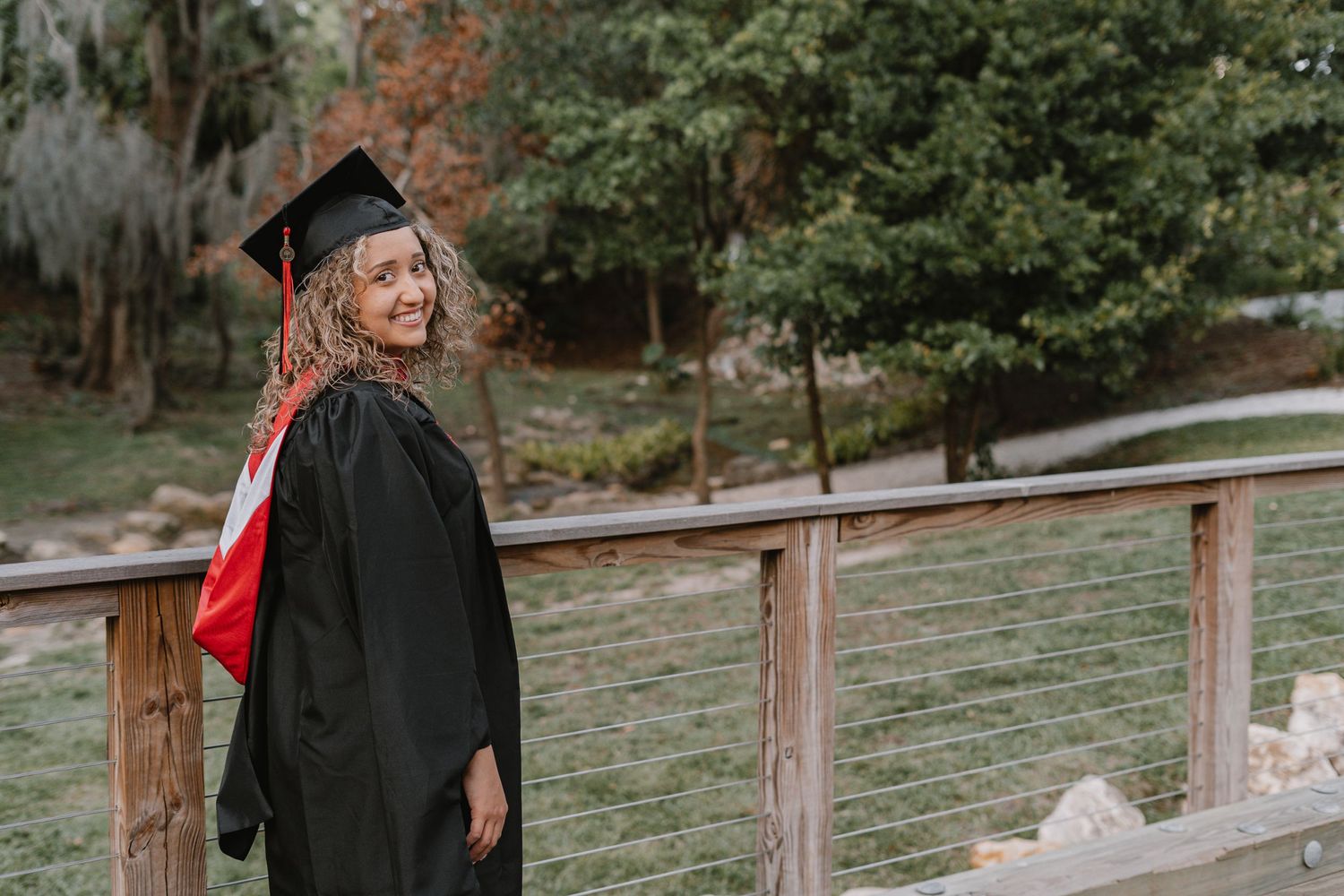 A graduate in black cap and gown with red stole stands smiling on a wooden deck outdoors.