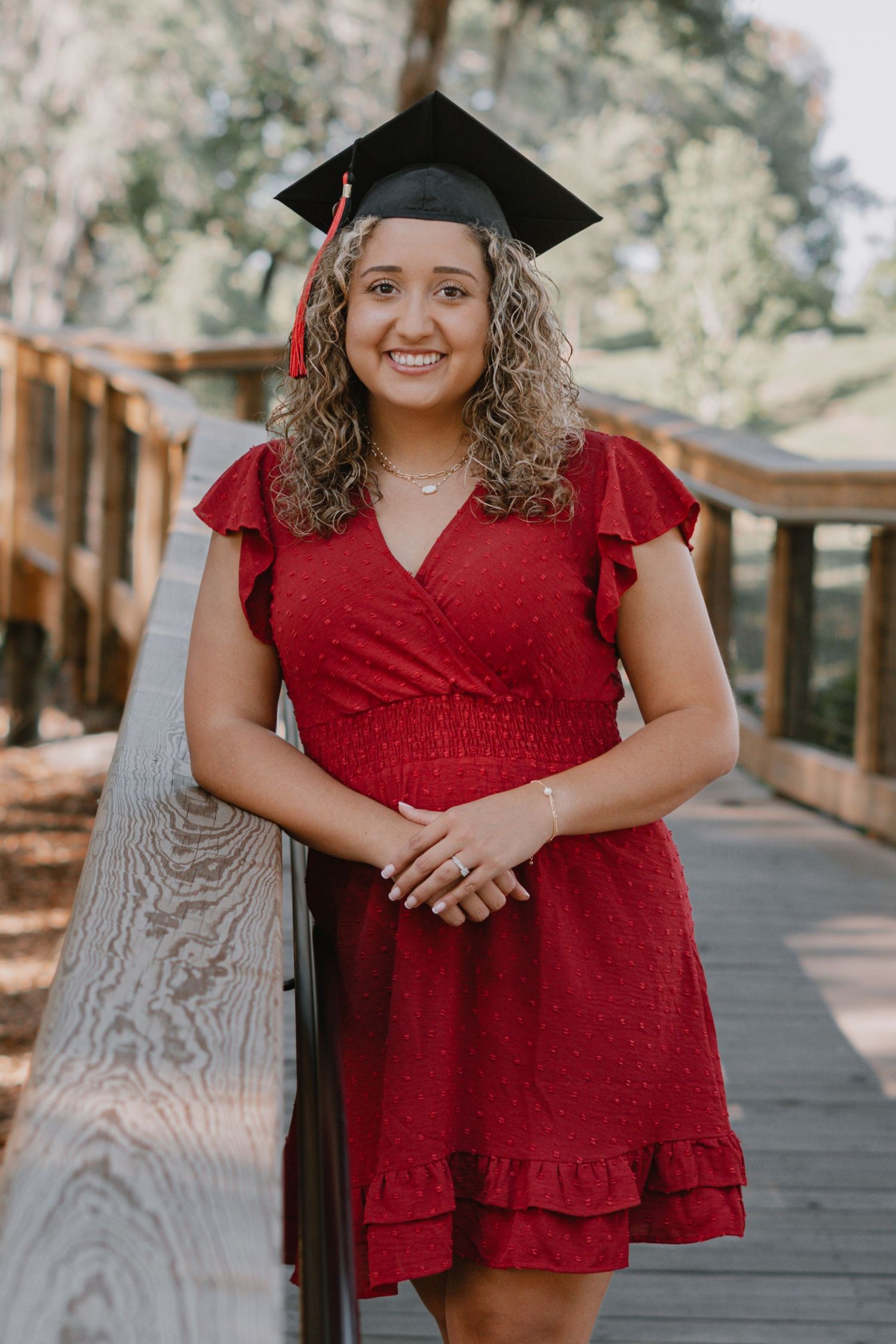 A graduate in a burgundy red dress and graduation cap poses on a wooden boardwalk outdoors.
