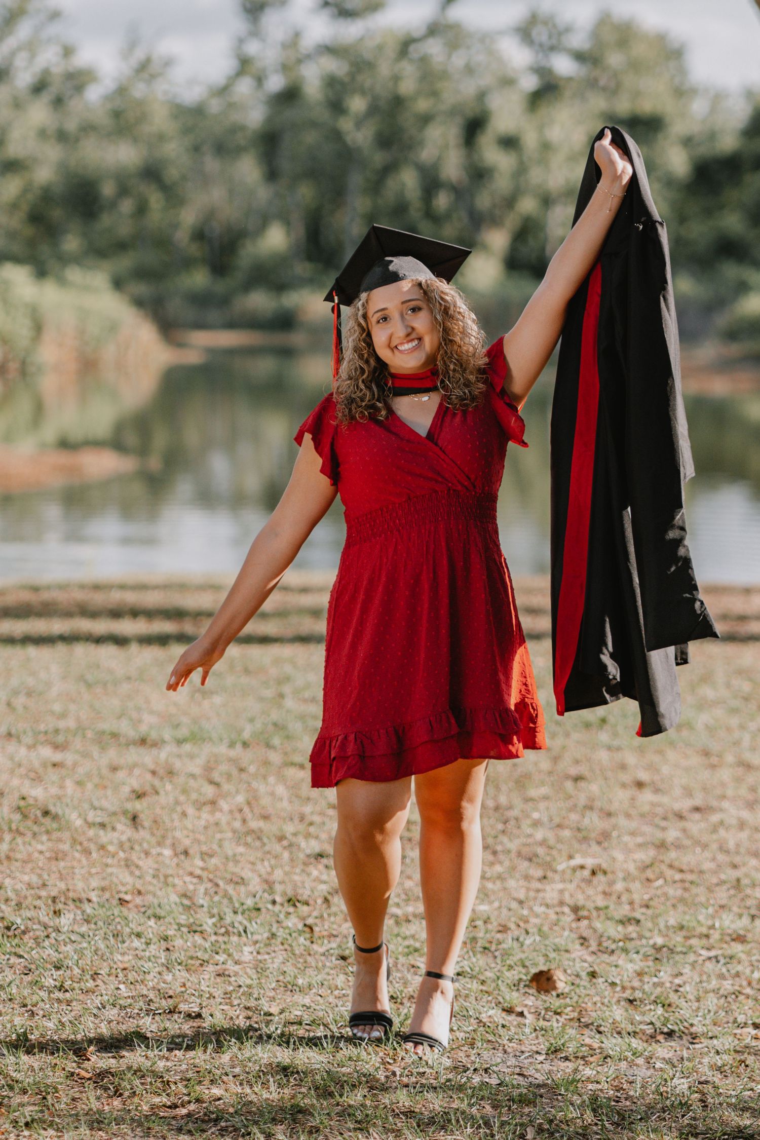 Graduate in red dress celebrates with cap and gown outdoors by a pond on a sunny day.